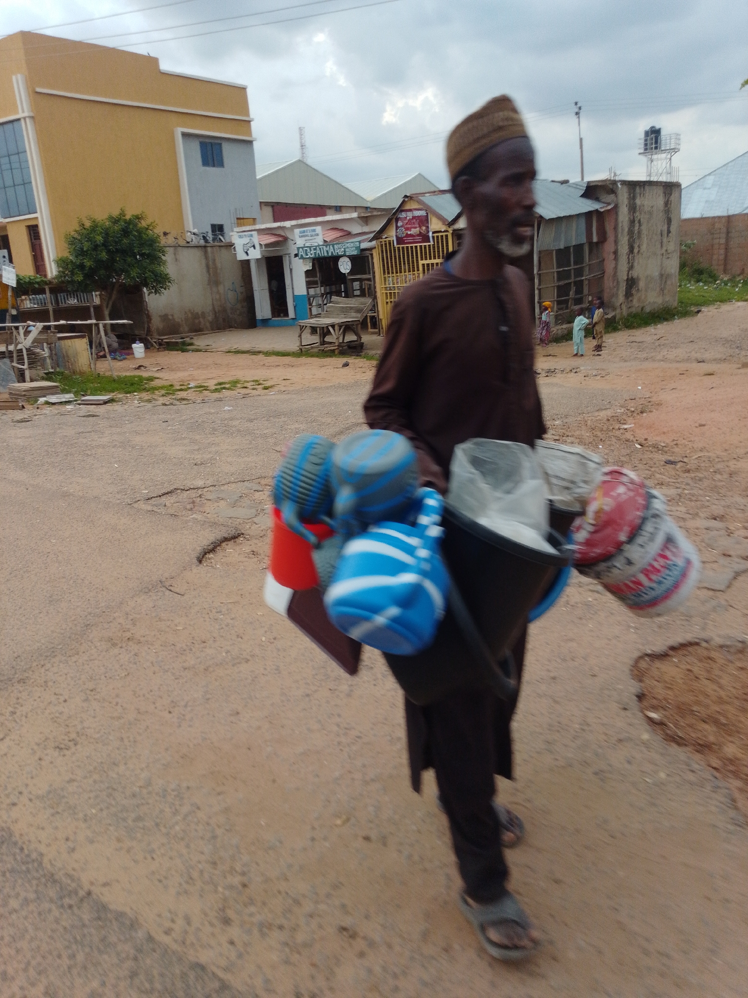 A man wearing a brown traditional outfit and a brown cap walks on a dusty road in Gombe, Nigeria. He carries a large black bucket filled with white sacks, and other containers are attached to his left side. These containers are red and white with some lettering visible and a blue and white patterned cover. In the background, several buildings are visible, including a yellow and grey multi-story building, a smaller building with a corrugated metal roof, and another structure with a similar roof. A few children are also visible in the background, standing near a building. The ground is a mixture of dirt and gravel, with patches of dry grass. The sky is overcast.