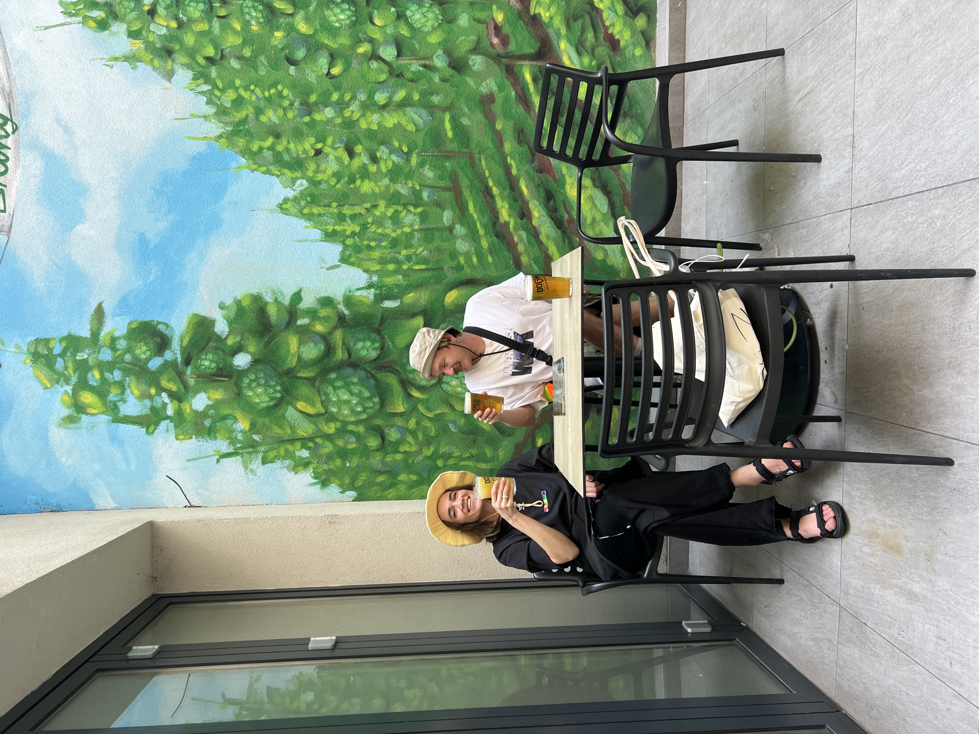 Two people are sitting at an outdoor table, each holding a glass of beer. A woman wearing a black t-shirt, black pants, and a straw hat is seated on the lower right. A man wearing a white t-shirt and a beige bucket hat is seated across the table from her. Behind them is a painted mural of hop vines on a building wall. The location is Dogma Brewery in Belgrade, Serbia. Two black chairs are visible at the table. A white tote bag is hanging on the chair near the man. The ground is tiled. A large window with a gray frame is visible on the lower left.
