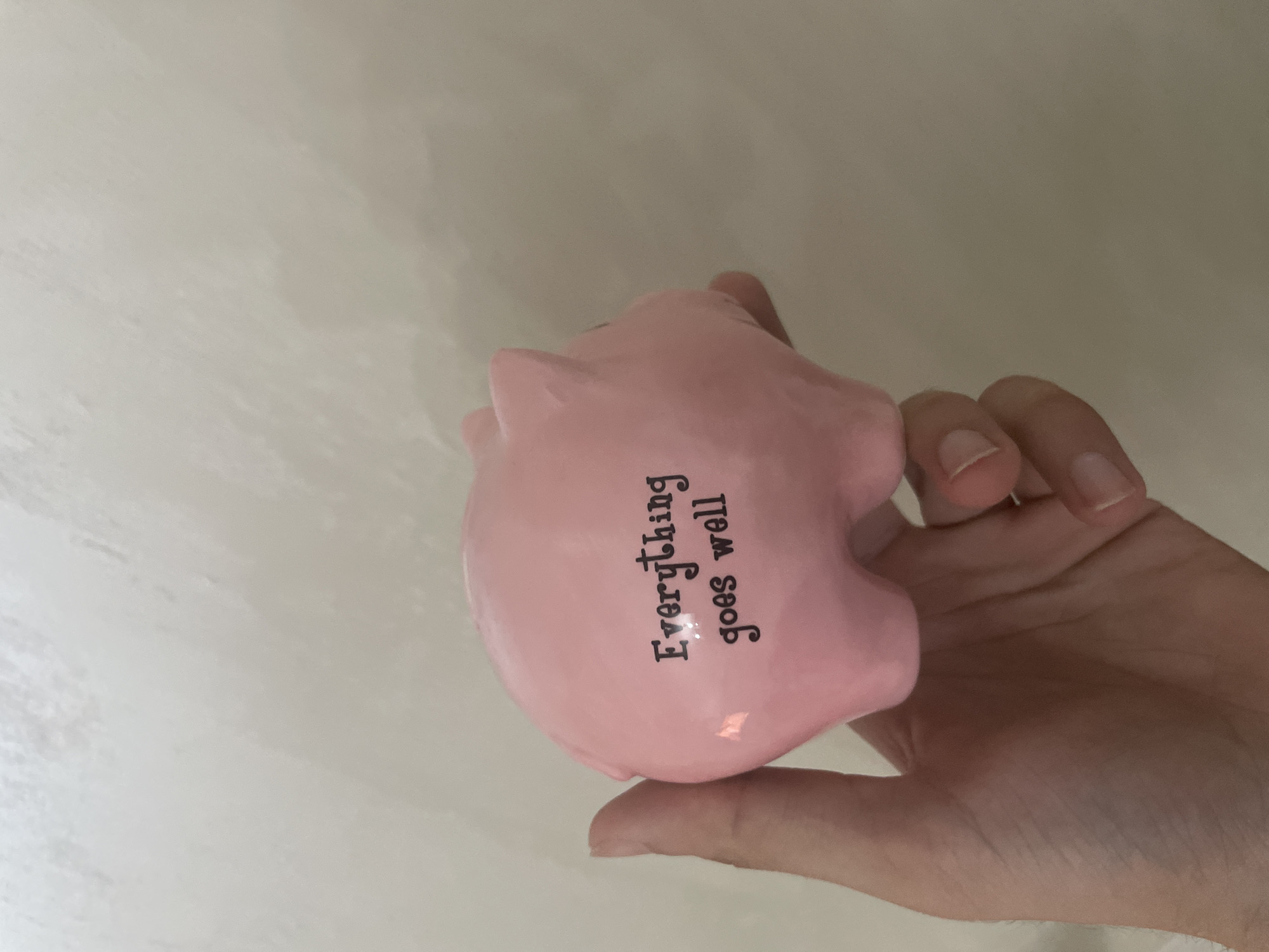 A human hand holds a pink ceramic piggy bank, which has the phrase "Everything goes well" inscribed in black block and lowercase letters on its side. The hand is positioned underneath and partially around the piggy bank, with visible fingers and parts of the palm. The piggy bank features molded details such as small ears, a snout, and four short legs. The background consists of a plain, light, neutral-colored surface, possibly a wall, exhibiting subtle texture and variations in shade. The scene is evenly lit, with no harsh shadows. The depicted scene, stated to be located in the city of Ihtiman, Bulgaria, does not contain specific environmental details within the image itself visually confirming this geographical context.