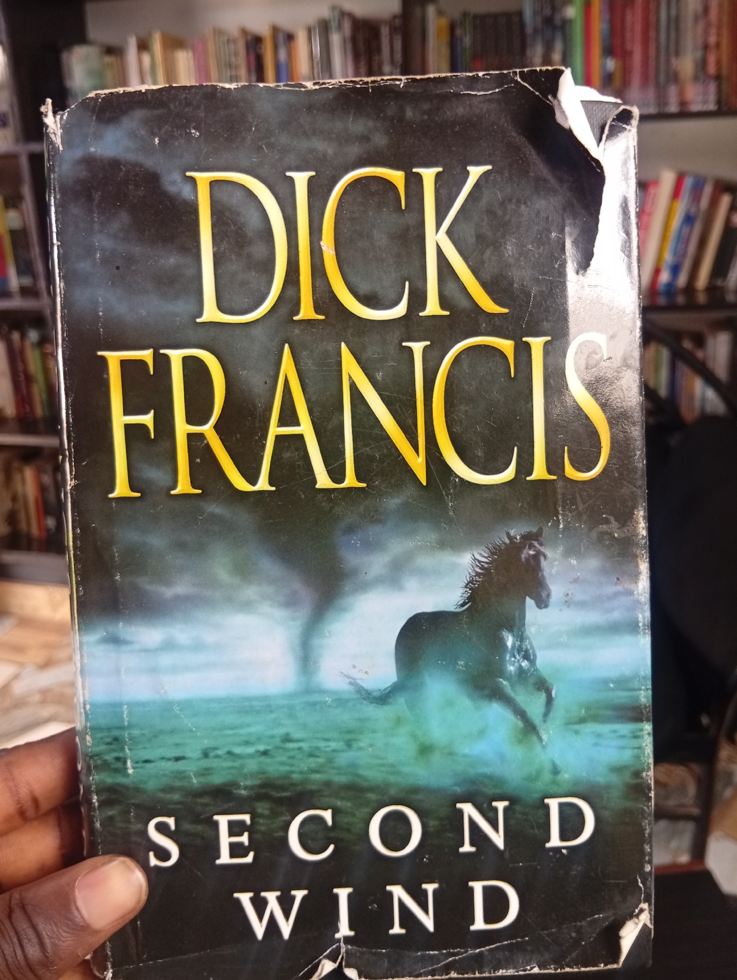 A close-up, eye-level shot captures the front cover of a book titled "SECOND WIND" by DICK FRANCIS. The cover features a dark, atmospheric background with a swirling vortex or tornado in the distance over a greenish, rippling body of water. In the foreground, a dark horse with a wet mane is depicted mid-stride in the water, appearing to break through the surface. The book cover itself shows signs of wear and tear, particularly at the top edge, with some peeling and scuff marks. A person's hand and thumb, belonging to someone with dark skin, are visible on the left side of the frame, holding the book. The background suggests the book is on display or being held in a location with bookshelves filled with other books, implying a library or bookstore setting. The scene appears to be indoors, with ambient lighting. There is no specific indication of the city of Jos, Nigeria within the image content itself.