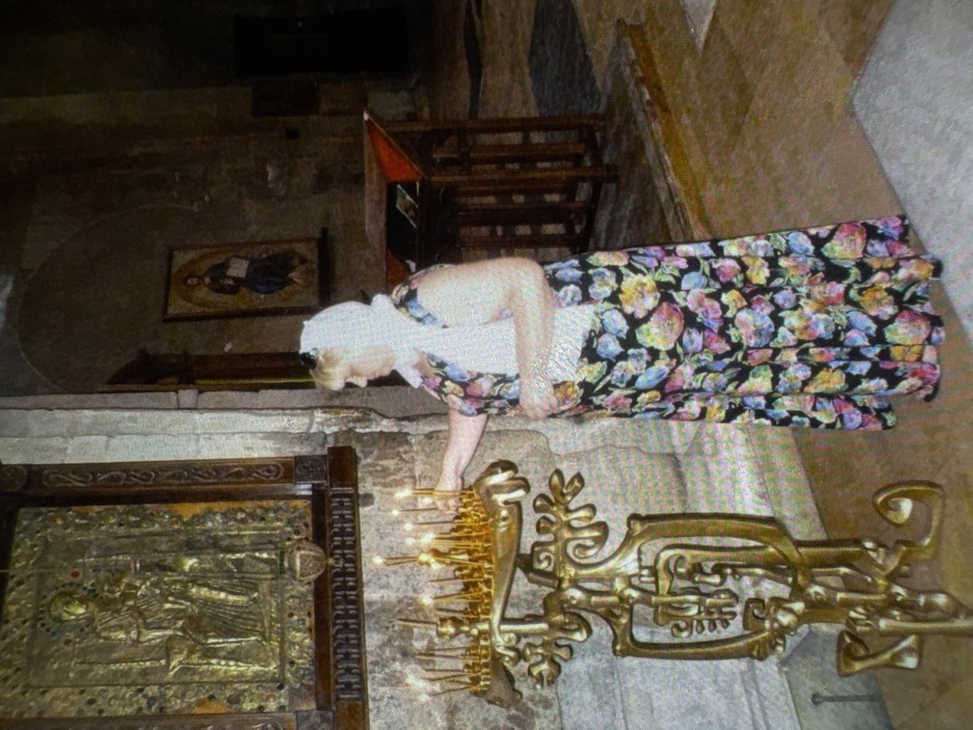 A middle-aged woman with blonde hair, wearing a white headscarf and a floral dress, stands in a dimly lit room, likely a church, in Batumi, Georgia. She is reaching out to light a candle from a large, ornate brass candelabra. The candelabra holds numerous lit candles, casting a warm glow. Behind the woman, a large, framed religious icon is mounted on a stone wall. To the right, a wooden lectern with books is visible. The floor appears to be made of stone tiles. The overall atmosphere is one of solemnity and devotion.