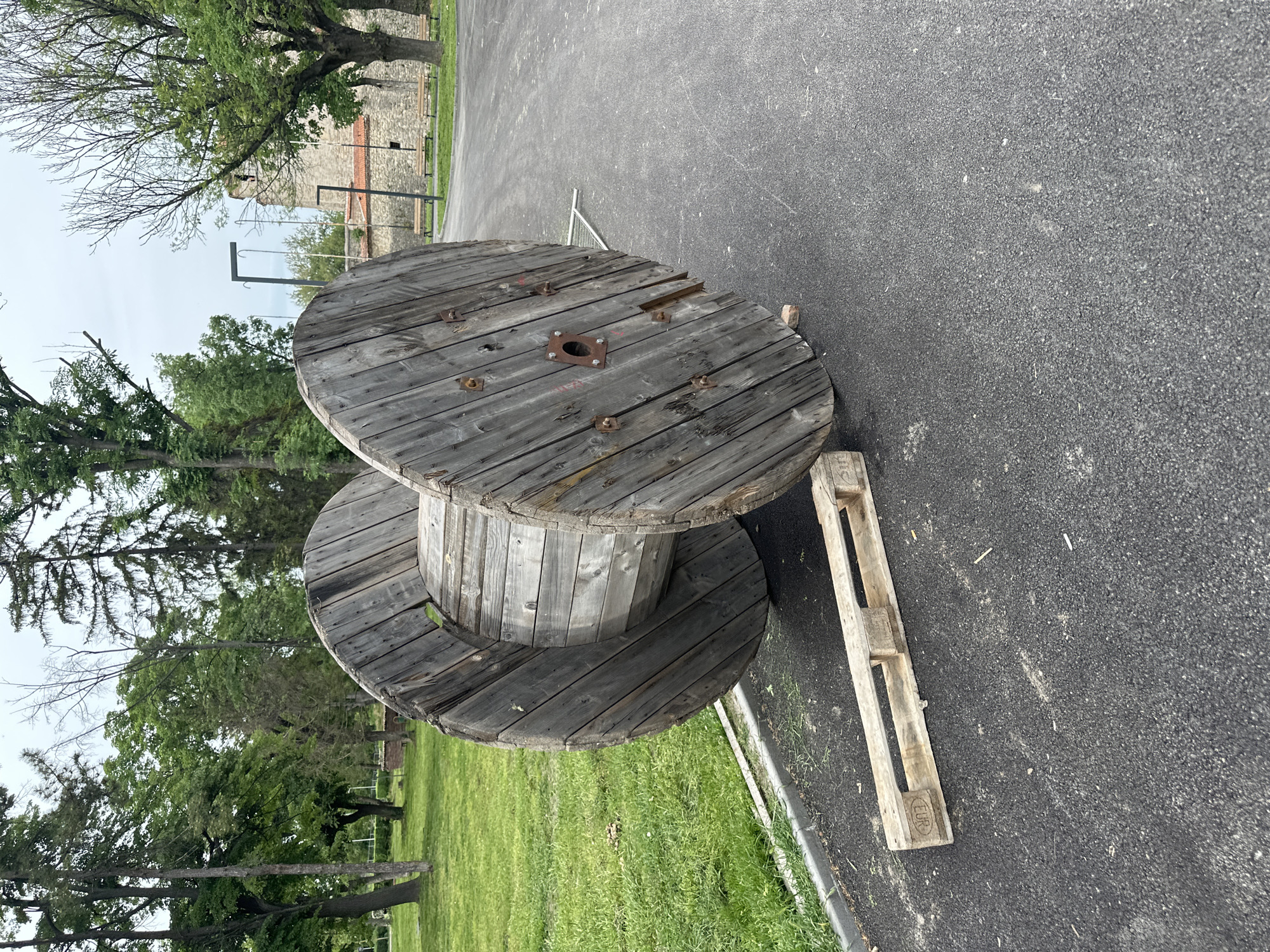 The image shows a large wooden cable reel and a wooden pallet lying on an asphalt surface next to a grassy area. The cable reel is weathered and gray, with visible bolts and a central metal plate. The pallet is light-colored wood and has visible markings. The background includes trees, grass, and a distant building with stone and brickwork. The location is Vidin, Bulgaria.