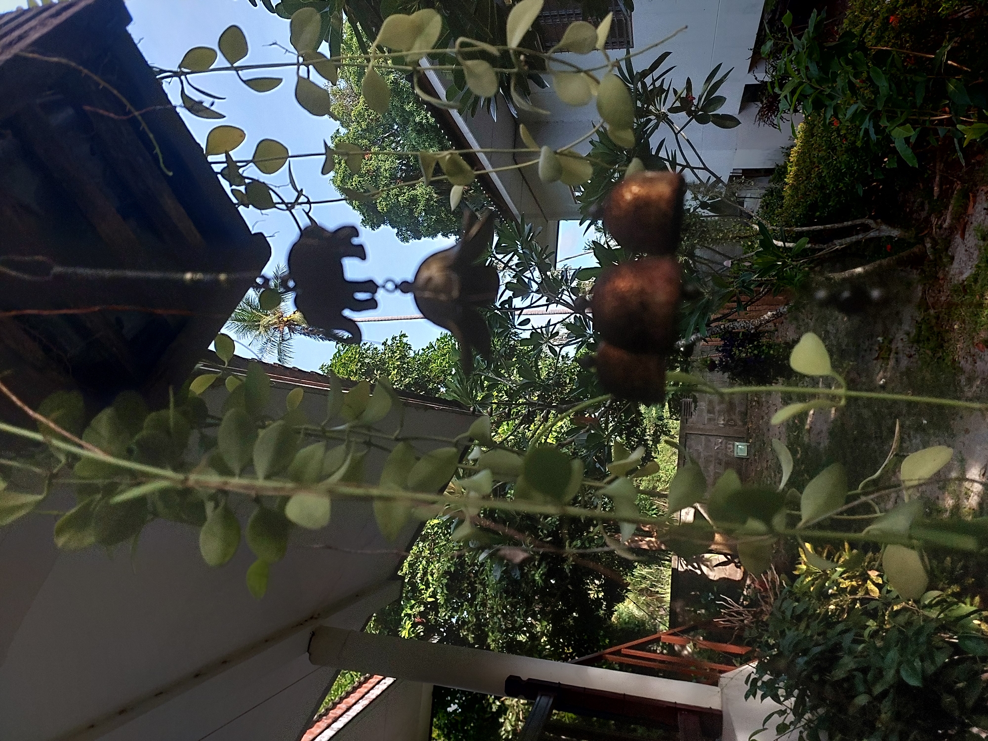 The image depicts a garden scene with a hanging wind chime in the foreground. The wind chime features an elephant silhouette and three copper bells. In the background, a white building with a tiled roof is visible, along with lush green trees and foliage. The sky is clear and blue. The location is identified as Baan Mae Nam, Thailand. There are no people visible in the image. The scene appears to be a tranquil outdoor setting.