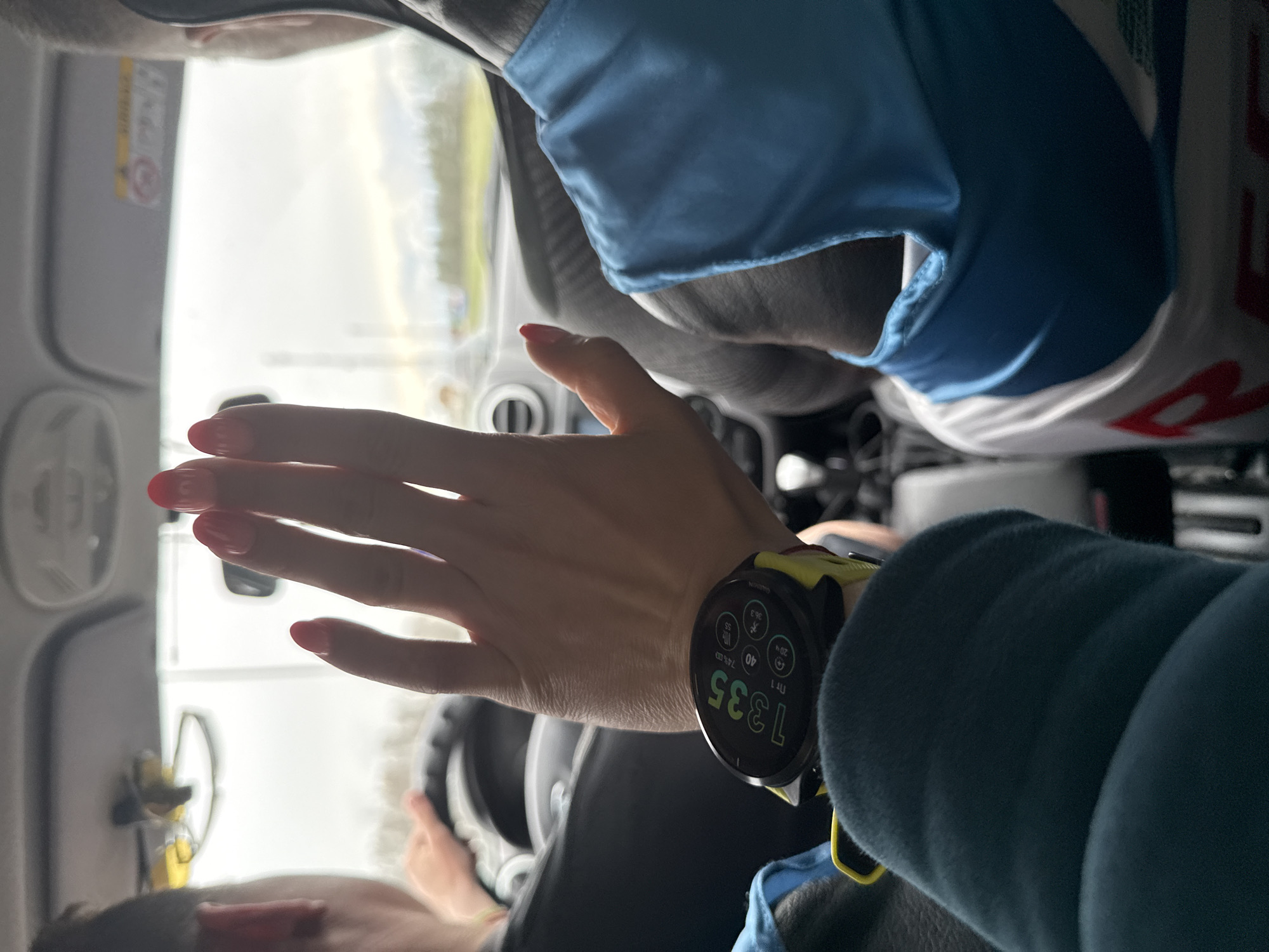 The image, taken from a first-person perspective inside a car in Bol'shie Nyrsi, Russia, centers on a person's arm and a prominent smartwatch. The arm, with red nail polish, is extended slightly, showcasing a yellow-banded Garmin smartwatch. The watch face displays the time as 13:35, along with "Пт" (Friday), "1 LU" (possibly 1st of the month), 74% battery, 40 steps, a heart rate of 55 bpm, a body battery/stress level of 36.3, and multiple notification icons.
In the background, a second individual, presumably the driver, is partially visible operating the steering wheel, wearing what appears to be a blue and white shirt. The foreground person wears a dark blue or green long-sleeved top. Through the car's windshield, a bright, possibly overcast, daytime scene is visible, indicating the car is on a road. Inside the car, parts of the dashboard and a sun visor with an "AIRBAG" warning are discernible. The overall scene depicts a moment during a car journey.
