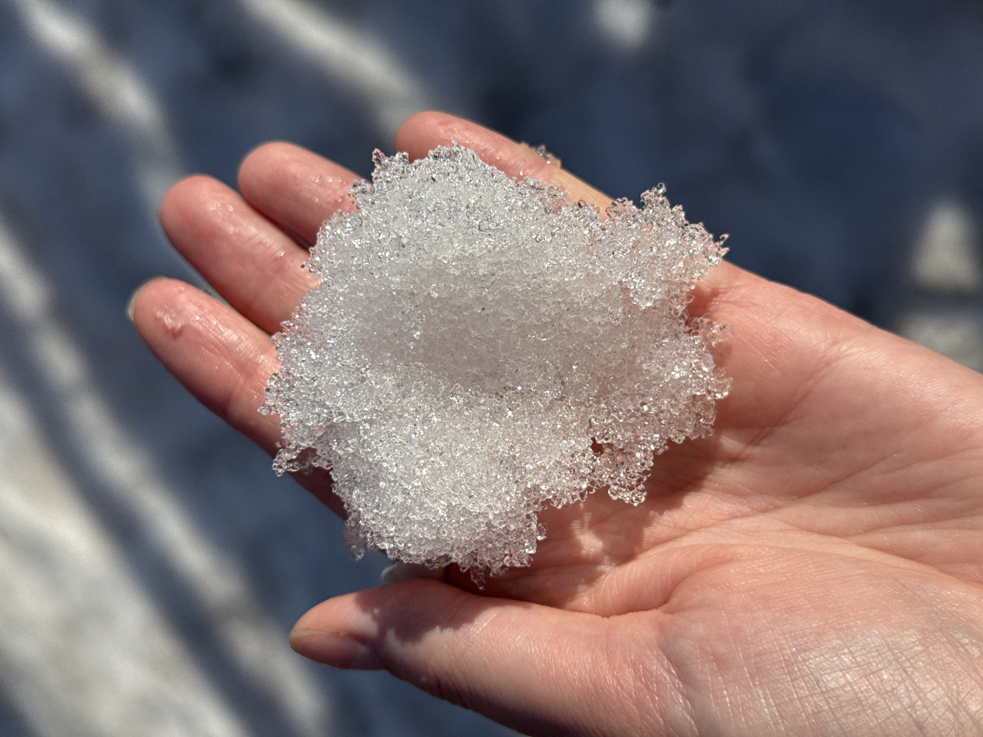 A person's open hand holds a clump of melting snow or ice crystals. The hand is pink-skinned, and appears slightly wet from the melting snow. The snow is a cluster of small, translucent, ice particles. The background is out of focus, but appears to be a snowy or icy surface, with shadows suggesting sunlight. There is no indication in the image as to it's location in Izhevsk, Russia.