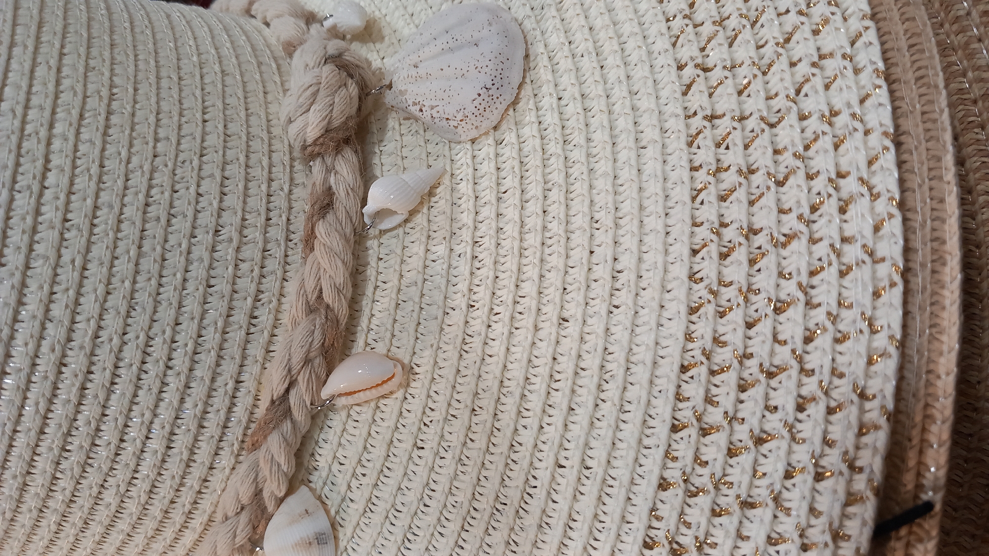 This image is a close-up shot of a straw hat. The hat is white and woven with a textured pattern. A thick, braided rope encircles the crown of the hat, and several small seashells are attached to the rope, dangling as embellishments. Some of these shells are white, while others have a light brown or tan color. In the lower portion of the image, there is a section of the hat that is woven with thin gold threads, creating a shimmering effect. The background is out of focus, but appears to be part of the same hat, suggesting it is a wide-brimmed style. There are no people, actions, or interactions visible. The image does not provide any information or visual cues about its location in the city of Kos, Greece.