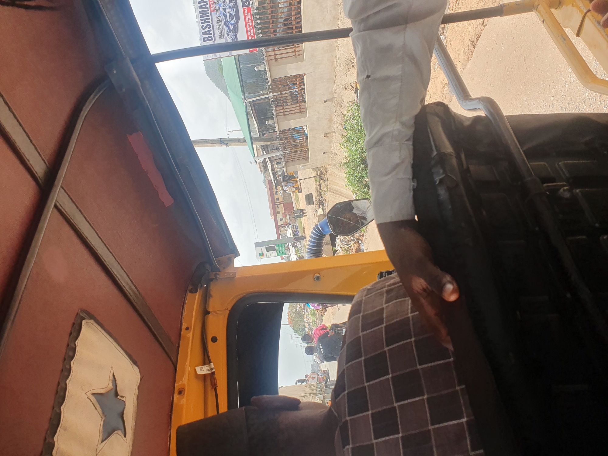 The image captures a tilted view from inside a yellow-framed commercial vehicle, likely a tricycle (Keke Napep), looking out onto a street scene. In the immediate foreground, the back of a person's head and shoulder, wearing a brown and white checkered shirt, is visible. Another person, dressed in a white long-sleeved shirt and light-colored trousers, has their arm and lower leg visible on the right side of the frame, holding onto part of the vehicle's structure.
The interior of the vehicle features a reddish-brown roof lining with a decorative star-shaped cutout and a piece of red tape. The yellow frame of the vehicle is prominent. Outside, a dusty street unfolds with several buildings, some appearing to be residential or commercial structures, possibly under construction. An electric pole stands tall, and patches of green vegetation are scattered along the roadside.
A large advertising signboard is clearly visible, reading: "BASHMAN DEALER BRAND NEW CARS, DIRECT BEL TLY OPPOSITE WADA PARK. E, LOKOJA, KOGI STATE." This text confirms the location as Lokoja, Kogi State, Nigeria, and indicates a car dealership. Other street activity, including people and vehicles, can be glimpsed through the side window and in the rearview mirror. A distinctive blue corrugated pipe or hose is also visible in the outdoor scene. The bright, clear sky indicates daytime, and the overall atmosphere suggests travel through a bustling urban or semi-urban area.