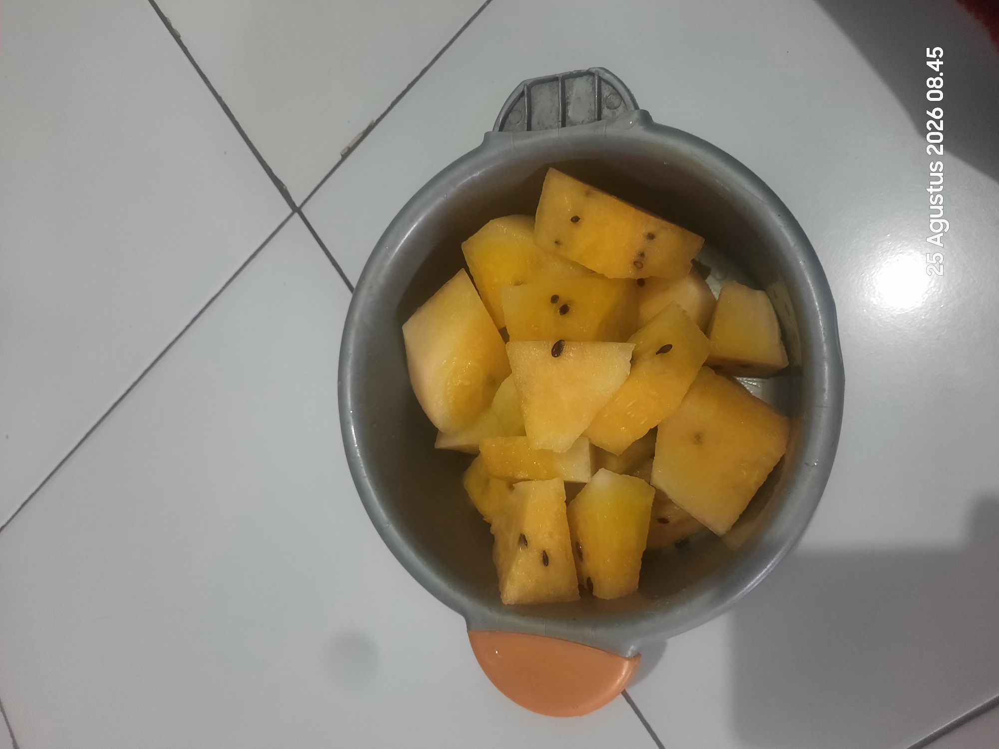 A bowl of chopped yellow watermelon sits on a tiled floor. The bowl is gray with an orange handle. The watermelon is cut into bite-sized pieces and has black seeds scattered throughout. The tiled floor is white with gray grout lines. A timestamp in the lower right corner of the image reads "25 Agustus 2026 08.45." The scene appears to be indoors, possibly in a kitchen or dining area, in Sukokerto, Indonesia.