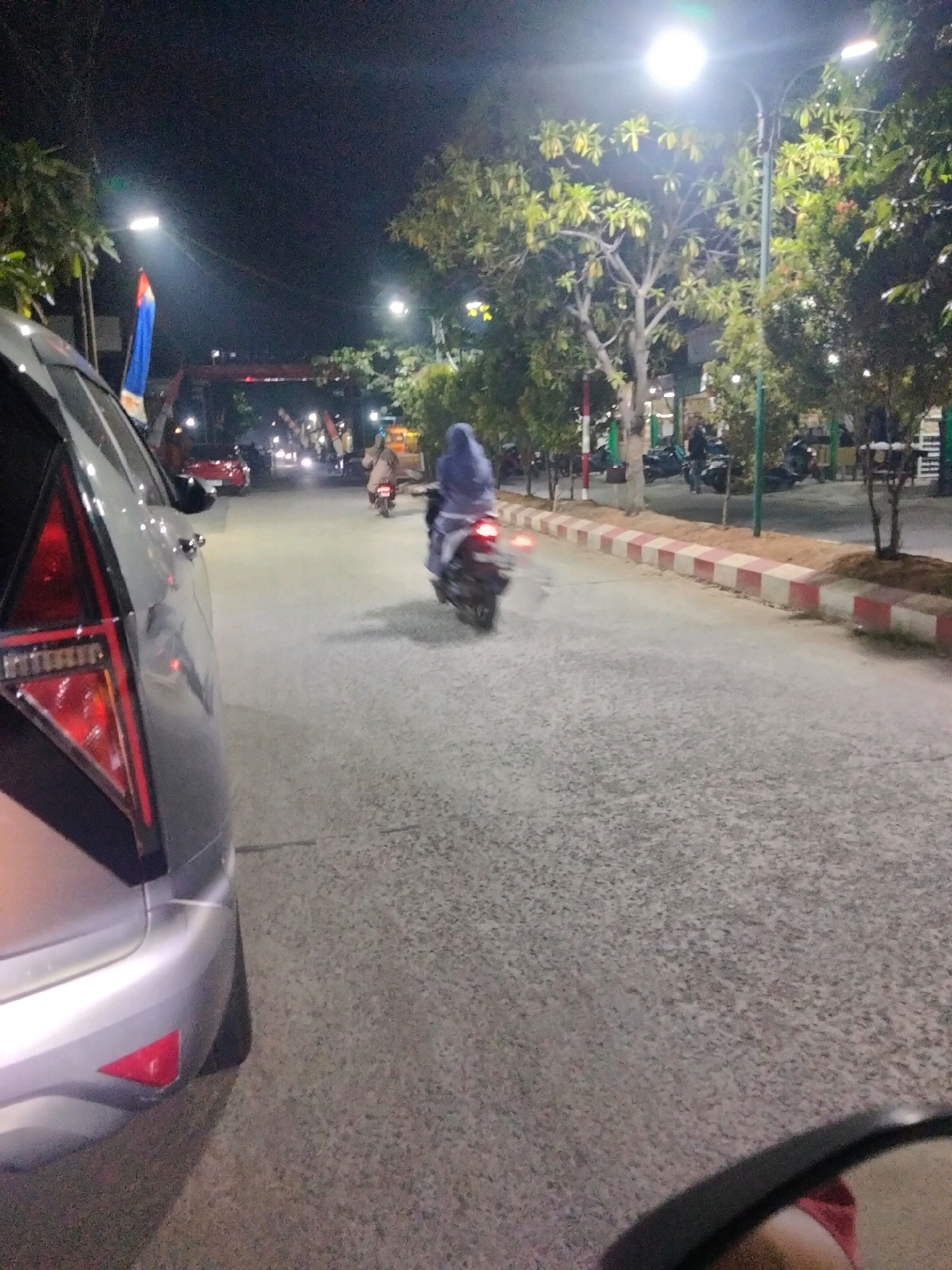The image depicts a street scene at night in Pisangan Jaya, Indonesia, under the illumination of streetlights. In the foreground, the rear section of a silver car is visible on the left. The road surface is asphalt, with a red and white checkered curb lining the right side. Further down the street, a person on a motorcycle is riding away from the camera, wearing a light-colored garment and a blue headscarf. Other motorcycles are parked along the right side of the road, near trees and buildings. In the background, several streetlights are on, and there appears to be some sort of overhead structure or bridge with lights. The atmosphere suggests a moderately busy urban environment at night.