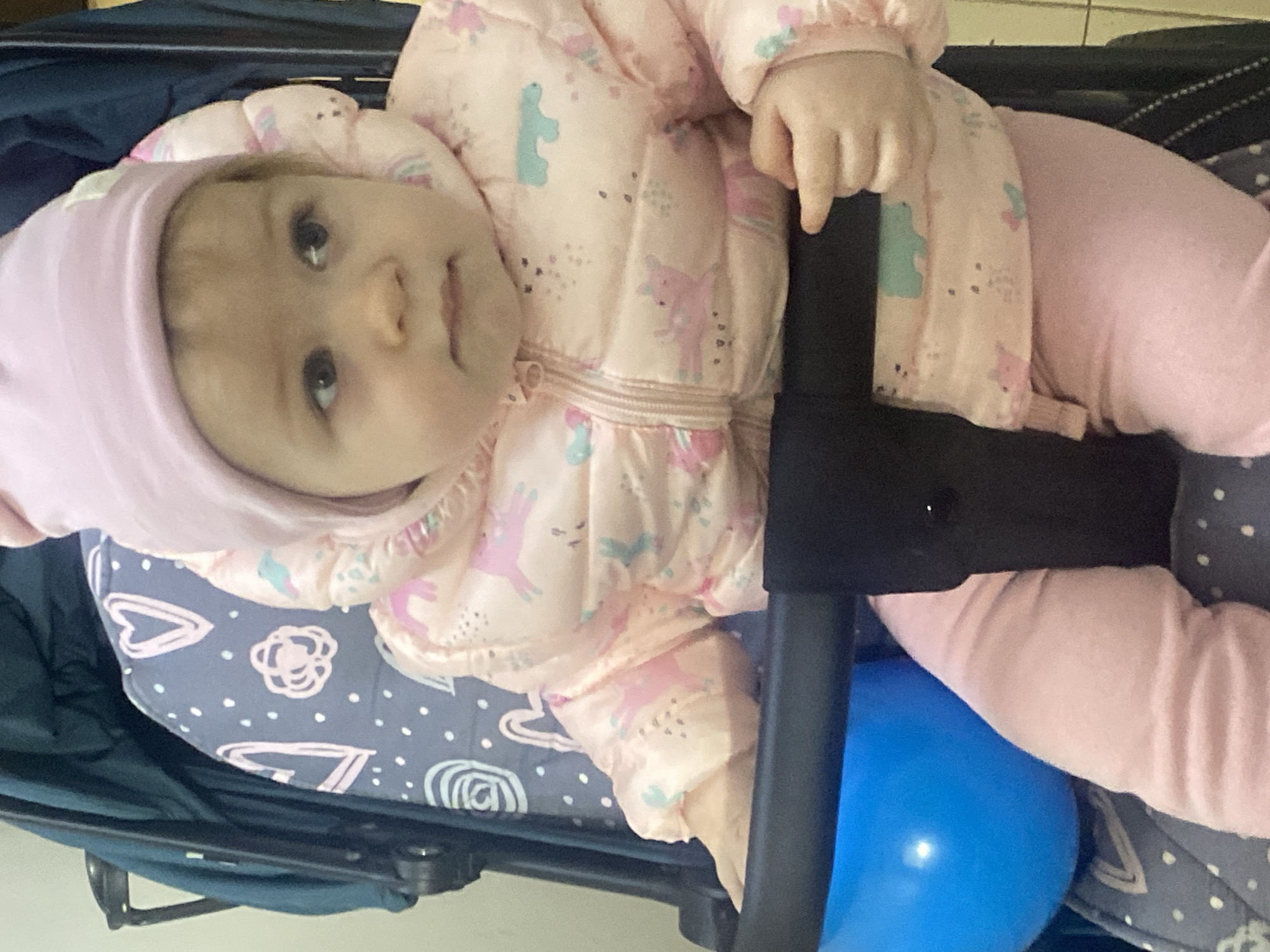 A baby wearing a pink hat and a pink jacket with unicorn and dinosaur patterns is secured in a stroller. The stroller has a blue and gray patterned seat liner with heart and flower designs. A bright blue object, possibly a toy or balloon, is visible near the baby's legs. The stroller is dark green or blue, and the image appears to be taken indoors with a plain wall in the background. Location details are unavailable.