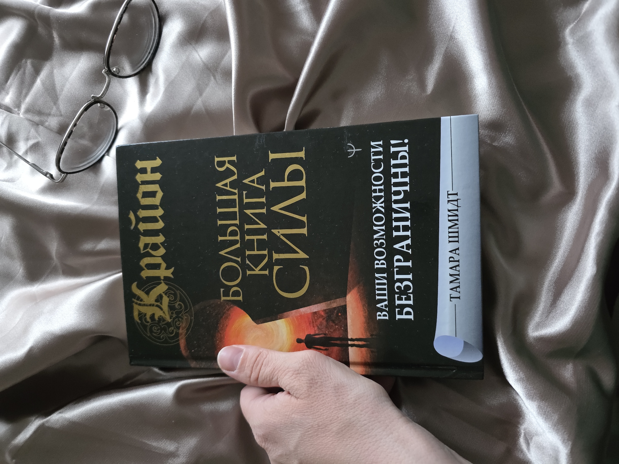 A person is holding a book titled "Большая книга силы" (The Big Book of Power) in Russian. The author is listed as "Тамара Шмидт" (Tamara Schmidt). A pair of eyeglasses rests beside the book on a wrinkled, satiny sheet. The setting appears to be indoors.