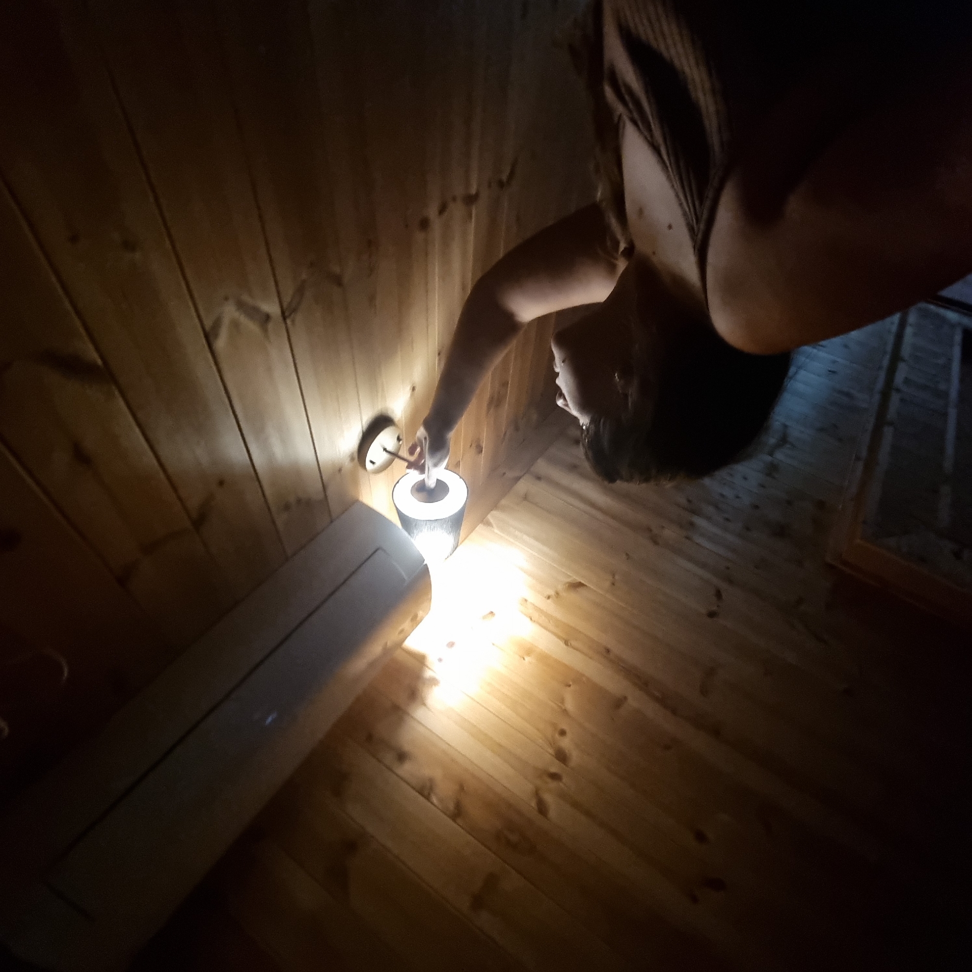 A woman with dark hair is reaching up to a wall-mounted light fixture in a room with wooden paneled walls and ceiling. The light fixture is a small, dark lampshade with a light bulb illuminating it. Above the light fixture, a white air conditioning unit is mounted on the wall. The room is dimly lit, with the brightest light coming from the lamp. The woman is wearing a dark tank top. The overall impression is of an interior shot, possibly in a residential setting.