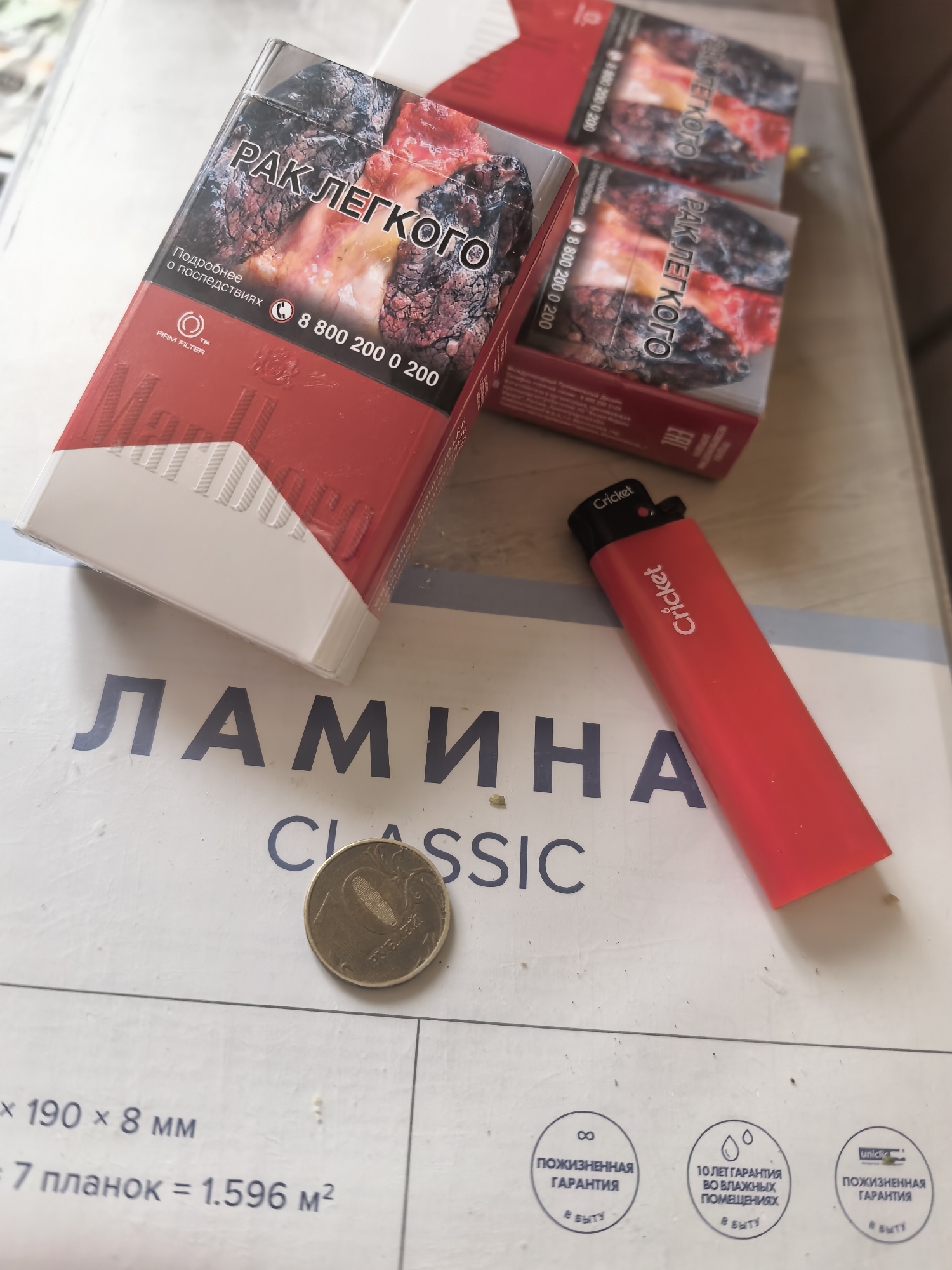 The image shows three packs of Marlboro cigarettes and a red Cricket lighter on top of a surface with text. The cigarette packs have warning labels in Russian, indicating "lung cancer". One pack is in focus and shows a graphic image of damaged lungs. The lighter is red and has the "Cricket" logo printed on it. The surface beneath the objects appears to be a printed material, possibly packaging or a poster, with text in Russian and some graphical elements. The text visible includes "ЛАМИНА" and "CLASSIC". There is also a coin visible on the printed surface, which appears to be a Russian 10 ruble coin. The lighting suggests an indoor environment, with a slight glare on the surfaces. The overall scene is a still life composition of smoking-related items.