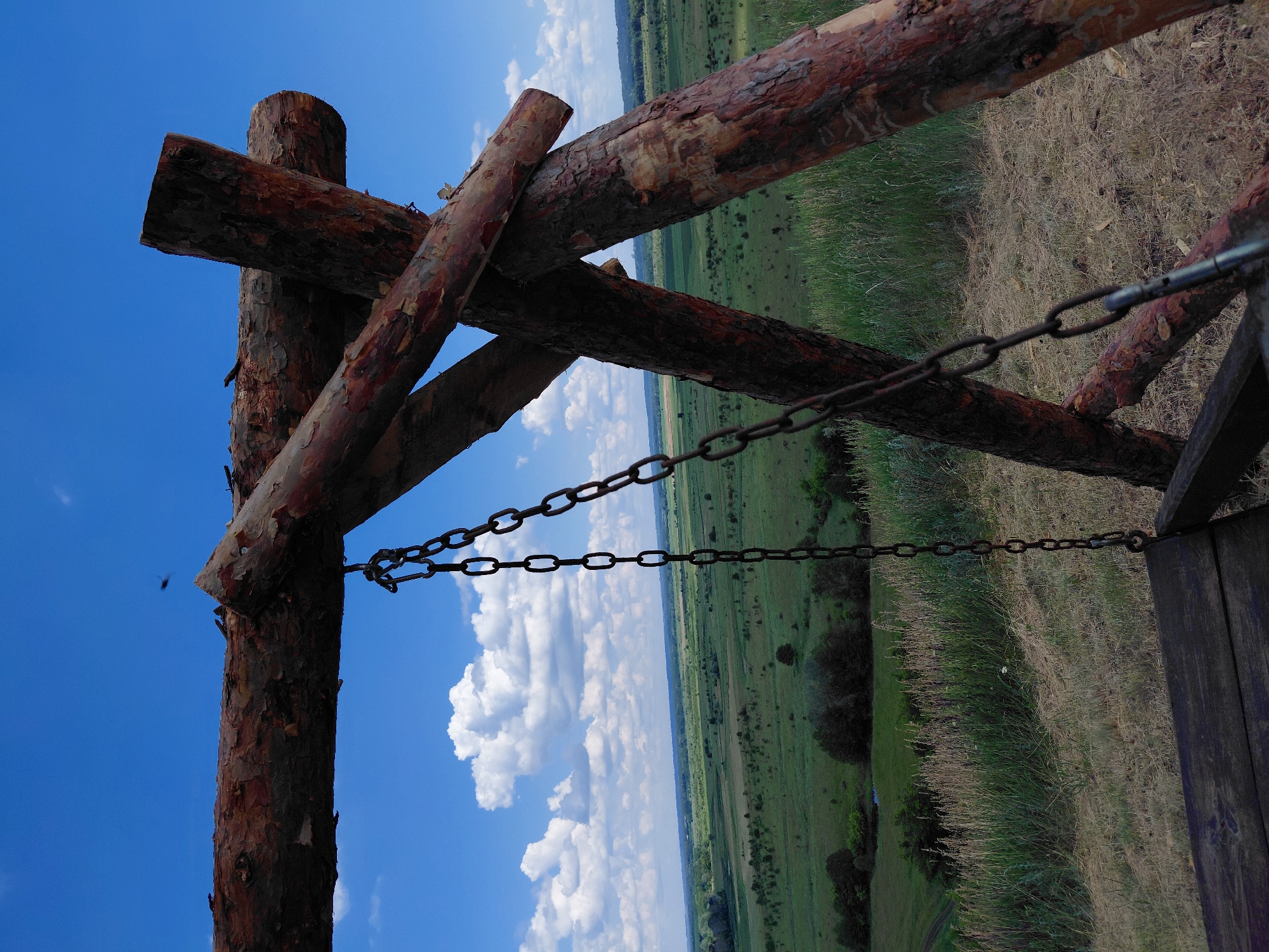 This is a close-up, eye-level, outdoor, vertical, medium shot photograph of a rustic wooden swing with a metal chain hanging in the foreground, set against a backdrop of a lush green valley under a bright blue sky dotted with white clouds. The swing is constructed from rough-hewn logs, with a horizontal beam forming the top support and two angled logs creating a triangular base on the right side of the frame. The metal chain hangs down on the left, attached to the horizontal beam and leading towards the bottom left corner where the seat of the swing is partially visible. The background reveals a wide expanse of rolling green fields, interspersed with scattered trees and bushes, indicative of a rural landscape. The sky is clear and blue with fluffy white cumulus clouds. The scene suggests a tranquil, scenic spot, possibly in a park or a natural setting. The location is identified as Ustyvytsia, Ukraine.