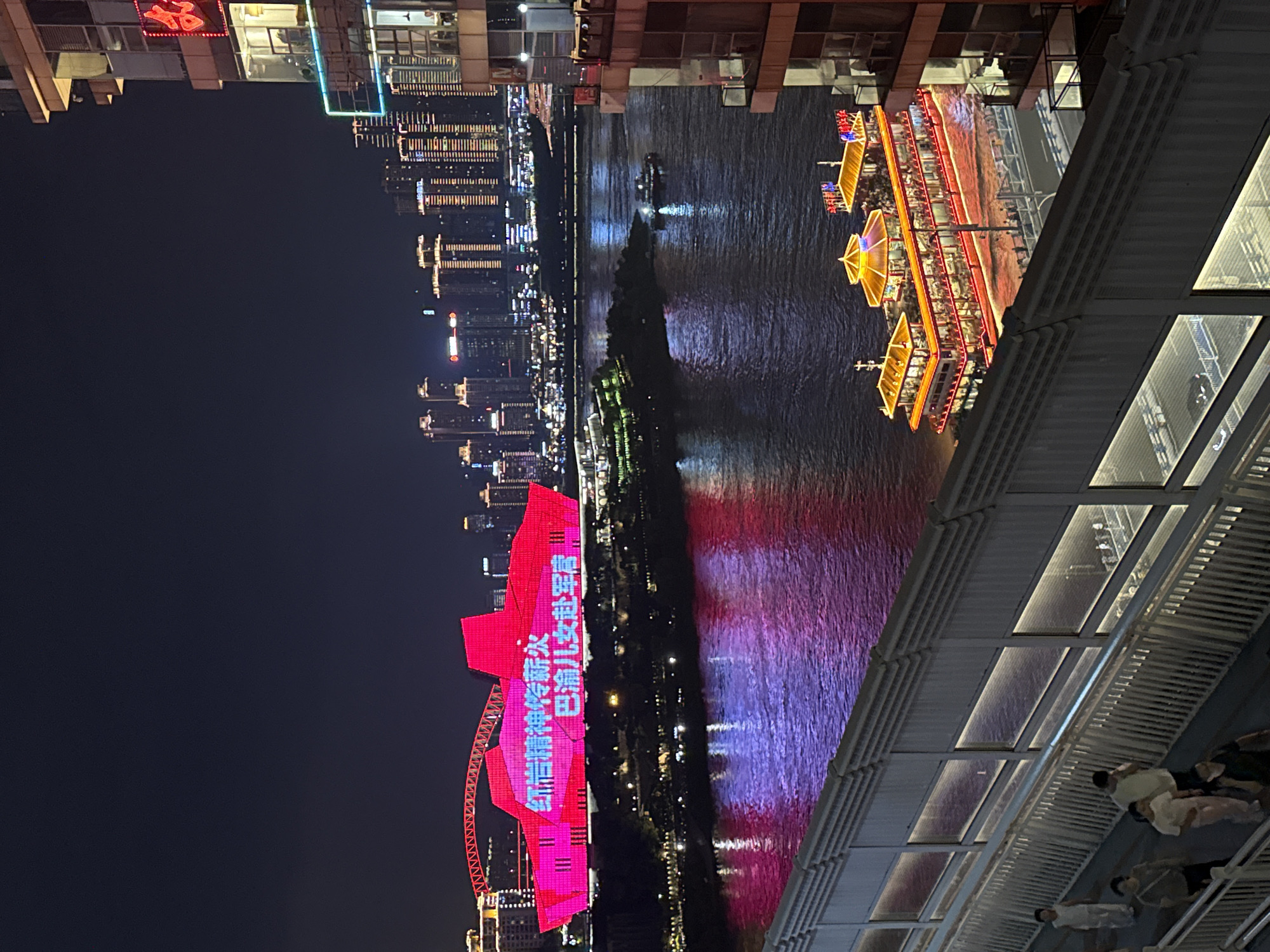 The image captures a night scene in the Yuzhong District of Chongqing, China, showcasing a vibrant urban landscape illuminated by artificial lights. In the foreground, a pedestrian walkway with white railings and glass panels overlooks a wide river. Several individuals are walking on the walkway.
Across the river, a prominent building with a distinctive, angular, red structure is brightly lit, displaying text in Chinese characters that appear to read "红岩精神薪火" and "巴渝儿女壮军营". This building is adjacent to a red, arched bridge. The river itself reflects the colorful lights from the buildings and the sky, creating a shimmering surface with hues of purple and red.
A large, illuminated tour boat with multiple decks and a traditional Chinese architectural style is prominently featured on the river, filled with people. In the background, a dense collection of modern skyscrapers with illuminated windows forms a striking city skyline. The overall scene conveys a lively and dynamic atmosphere of a bustling metropolis at night.