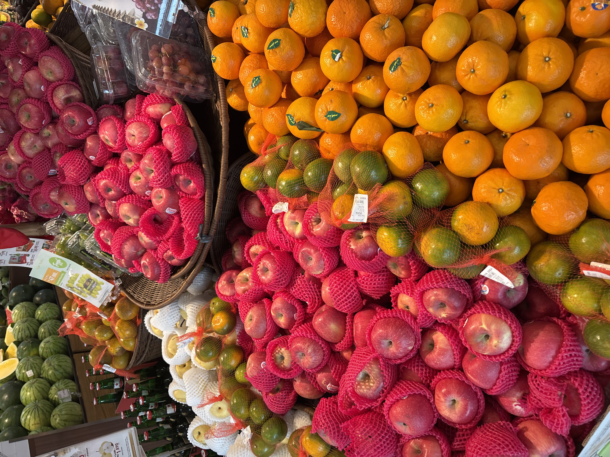 The image displays a vibrant fruit stall in Kuta Selatan, Indonesia, featuring an abundant array of fresh produce. In the foreground, a large pile of red apples, individually wrapped in pink foam netting, dominates the scene. To the right, a generous heap of bright orange tangerines, some with green leaves attached, are displayed, with a few mesh bags of green citrus fruits visible behind them. The middle ground showcases more red apples in similar netting, accompanied by a basket of golden-yellow fruits, likely pears or apples, and more mesh bags of green and yellow citrus. In the background, watermelons are visible, along with bottles of what appear to be beverages. The lighting suggests an indoor market or supermarket setting. No people are visible in the image.