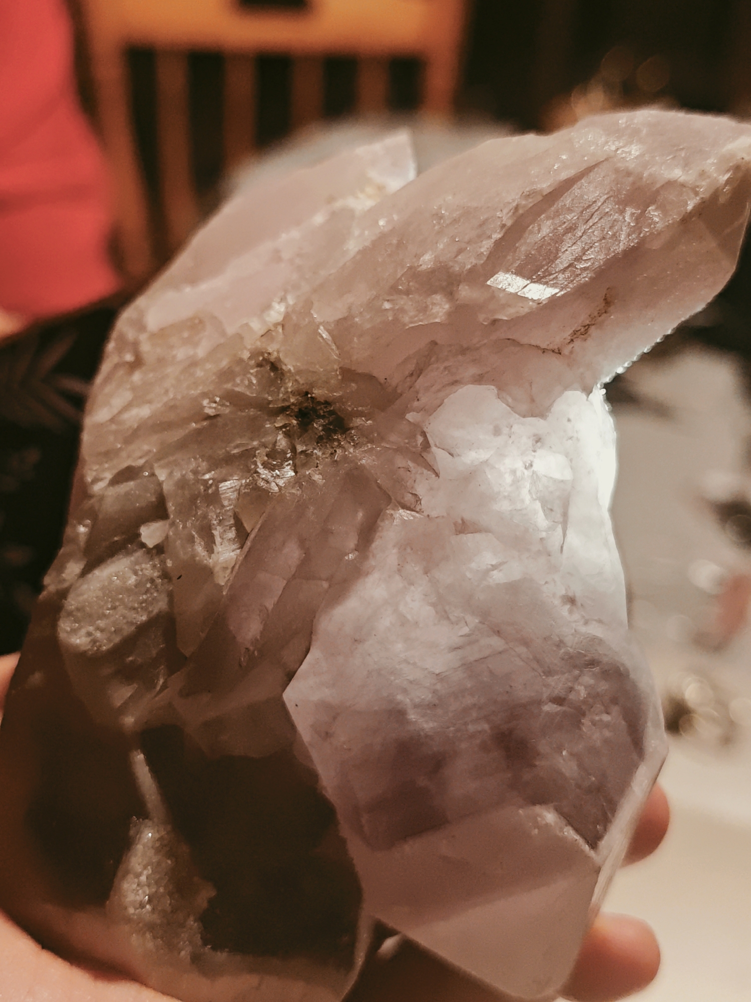 A person holds a large, multi-faceted quartz crystal. The crystal is translucent with a pale, smoky color, and has visible inclusions. The person's hand is visible at the bottom of the frame. The background includes a blurred view of a room with furniture and a person wearing a red shirt. The image does not contain any information indicating that it was taken in the city of Ильинское, Russia.