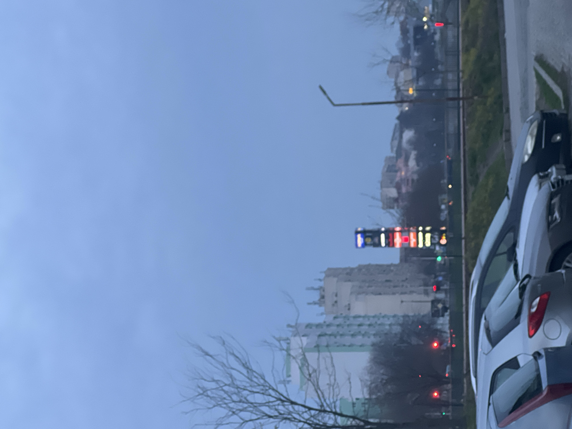 The image captures a cityscape under an overcast sky, likely taken during dawn or dusk. In the middle ground, a row of high-rise buildings is visible, some with white and green facades. A tall, thin structure stands out, possibly a tower or a large advertisement sign with multiple illuminated horizontal segments, displaying red, white, and blue colors. The road is on the right edge of the image, and several parked cars are arranged along the side of the street. Bare tree branches in the foreground suggest a winter or early spring season. The buildings suggest that the image may be taken somewhere in the city of Yambol, Bulgaria.