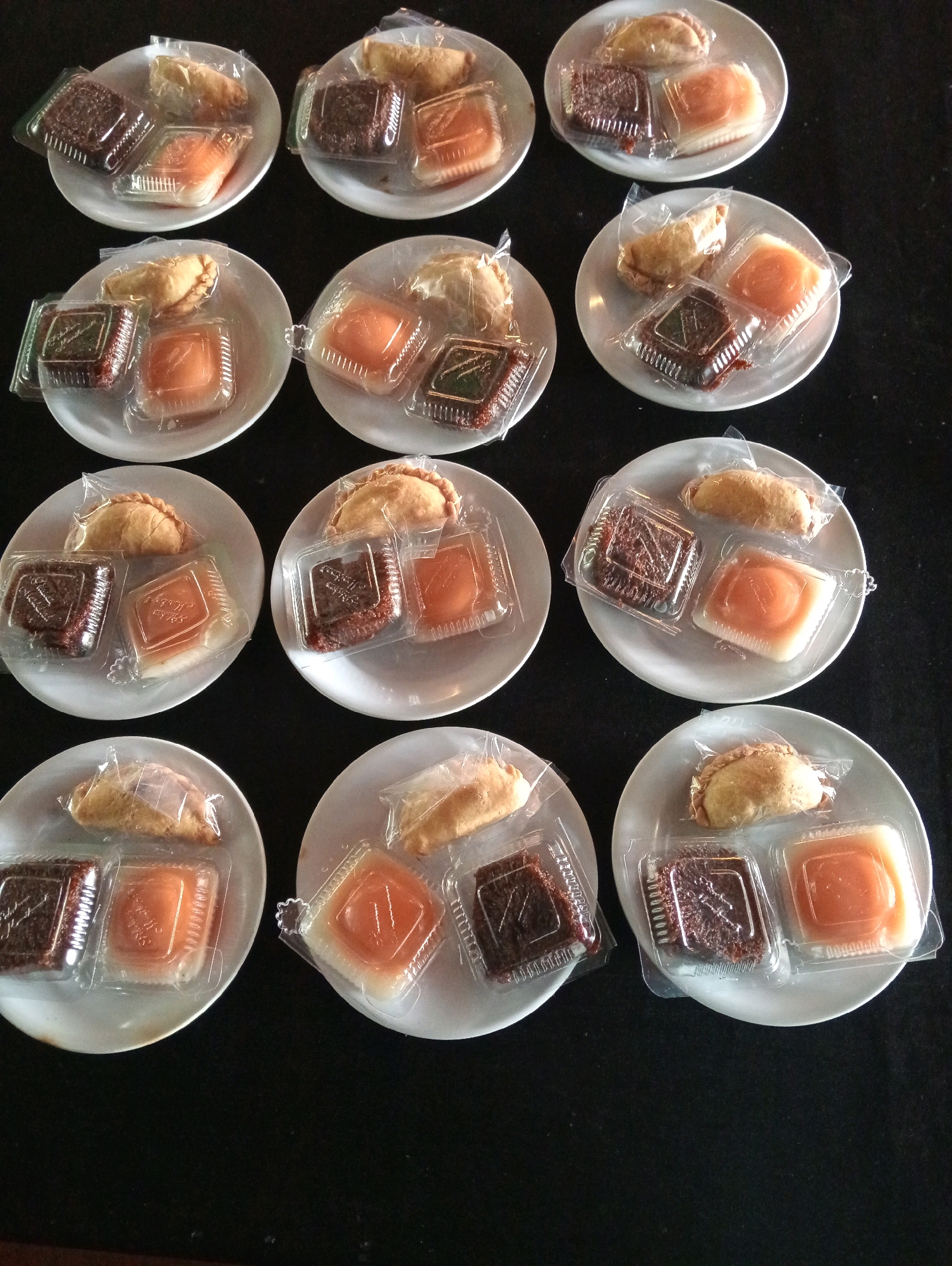 A close-up, overhead shot showcases an arrangement of twelve identical snack plates, likely set out for an event. Each white, circular plate holds three individually packaged items: a golden-brown pastry, a dark brown, dense confection in a clear plastic container, and a square of an orange-colored dessert in another clear plastic container. The plates are arranged in four rows of three, creating a visually uniform grid against a dark, textured background, possibly a tablecloth. The lighting is even, highlighting the textures and colors of the food. The overall scene suggests a prepared offering of sweets, possibly at a gathering or event in Tambak Rejo, Indonesia.