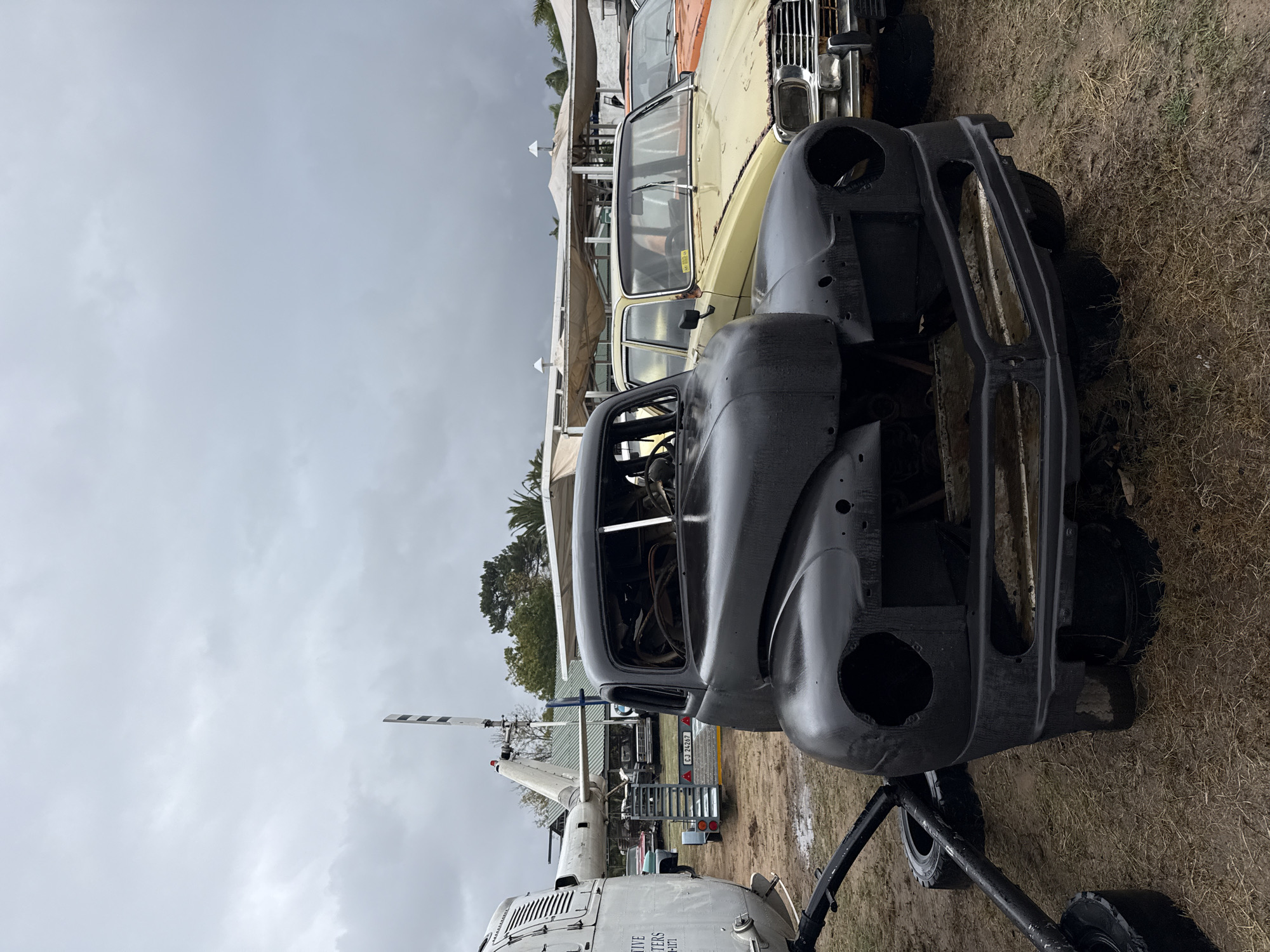 The image depicts a partly dismantled black car body parked on a dirt ground alongside other old vehicles in Kraaifontein, South Africa. In the foreground is part of a helicopter. Buildings and trees are visible in the background under a cloudy sky.