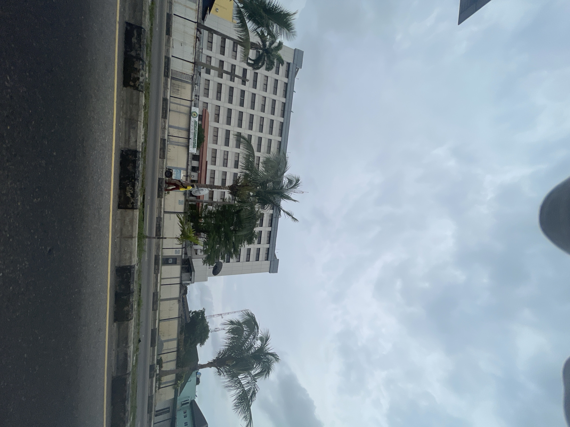 The scene shows a view of a street in Lagos, Nigeria, with a tall building and palm trees. Along the edge of the street, there is a sidewalk with a yellow line. A person wearing a yellow top and dark pants is walking on the sidewalk. The sky is overcast with gray clouds. There is a street with dark asphalt in the foreground.