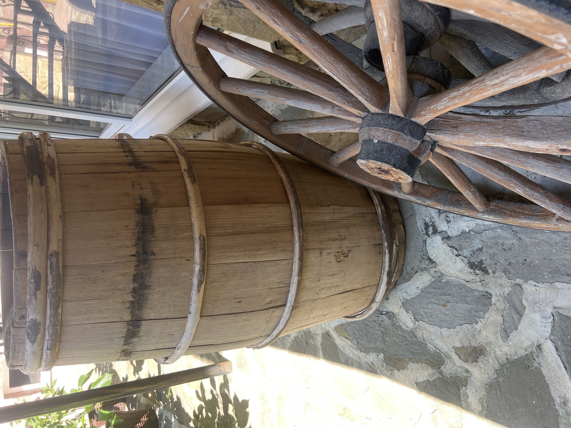 An outdoor scene features a large wooden barrel and a wooden wagon wheel leaning against a stone wall. The barrel is cylindrical with horizontal wooden staves held together by wooden hoops. The wagon wheel has a wooden rim and spokes. In the background, a window with white framing and sheer white curtains is visible, and beyond that, a metal staircase. The ground is paved with irregular stone slabs. The lighting suggests it is daytime, with sunlight casting shadows. The image is taken at a slightly low angle, looking up at the barrel and wheel. There are no people visible in the frame. The setting appears to be a rustic outdoor area, potentially in the city of Yaremche, Ukraine, given the context.