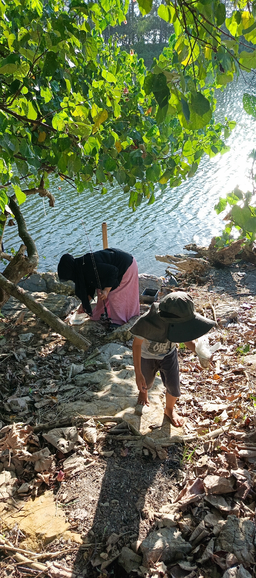 In Meunasahmesjid, Indonesia, two individuals are engaged in fishing by a body of water. An adult, dressed in a black headscarf and a pink striped skirt, is bent over near the water's edge, holding a fishing rod. To their right, a child wearing a wide-brimmed hat and shorts stands, holding a plastic bag. The ground is covered in rocks and fallen leaves, with tree branches and foliage overhead casting shadows. The water reflects the sunlight, creating a shimmering surface.