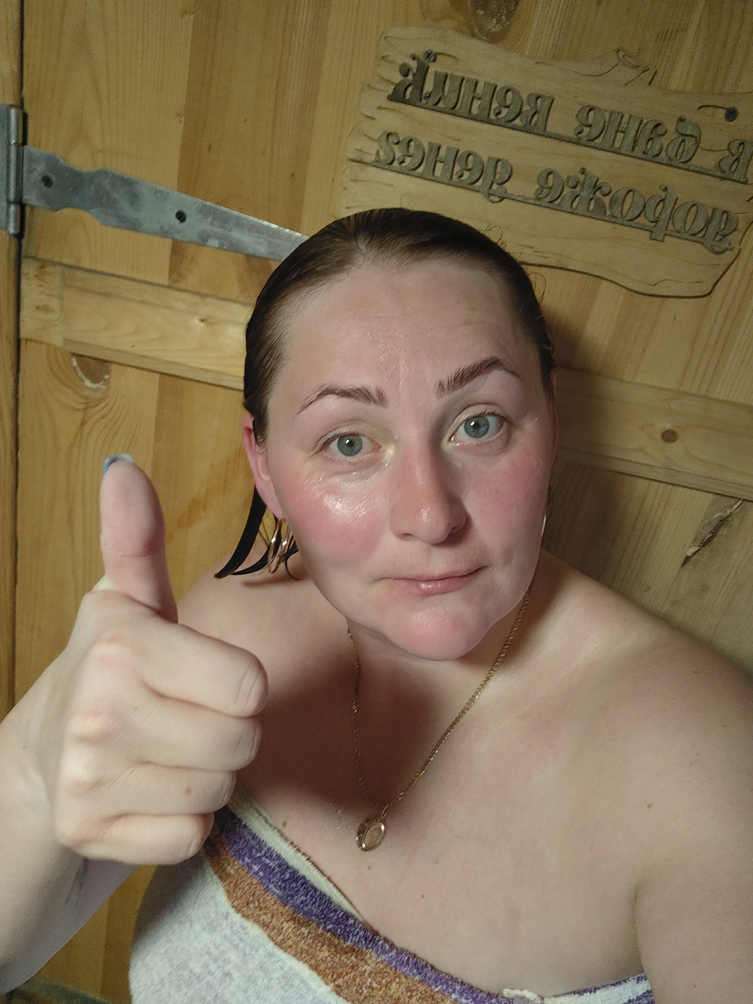 A woman with wet hair and reddened skin gives a thumbs-up gesture, potentially indicating approval or satisfaction. She is wearing a gold necklace and earrings, and a striped towel is draped around her upper body. The background consists of wooden planks, suggesting she is in a sauna or similar wood-paneled room. There is a metal hinge visible in the upper left corner and a wooden sign with text in Cyrillic script in the upper right. The presence of the sign in Cyrillic script points to the location being in a Russian-speaking area, and based on information from the user, the location is Фоки, Russia.