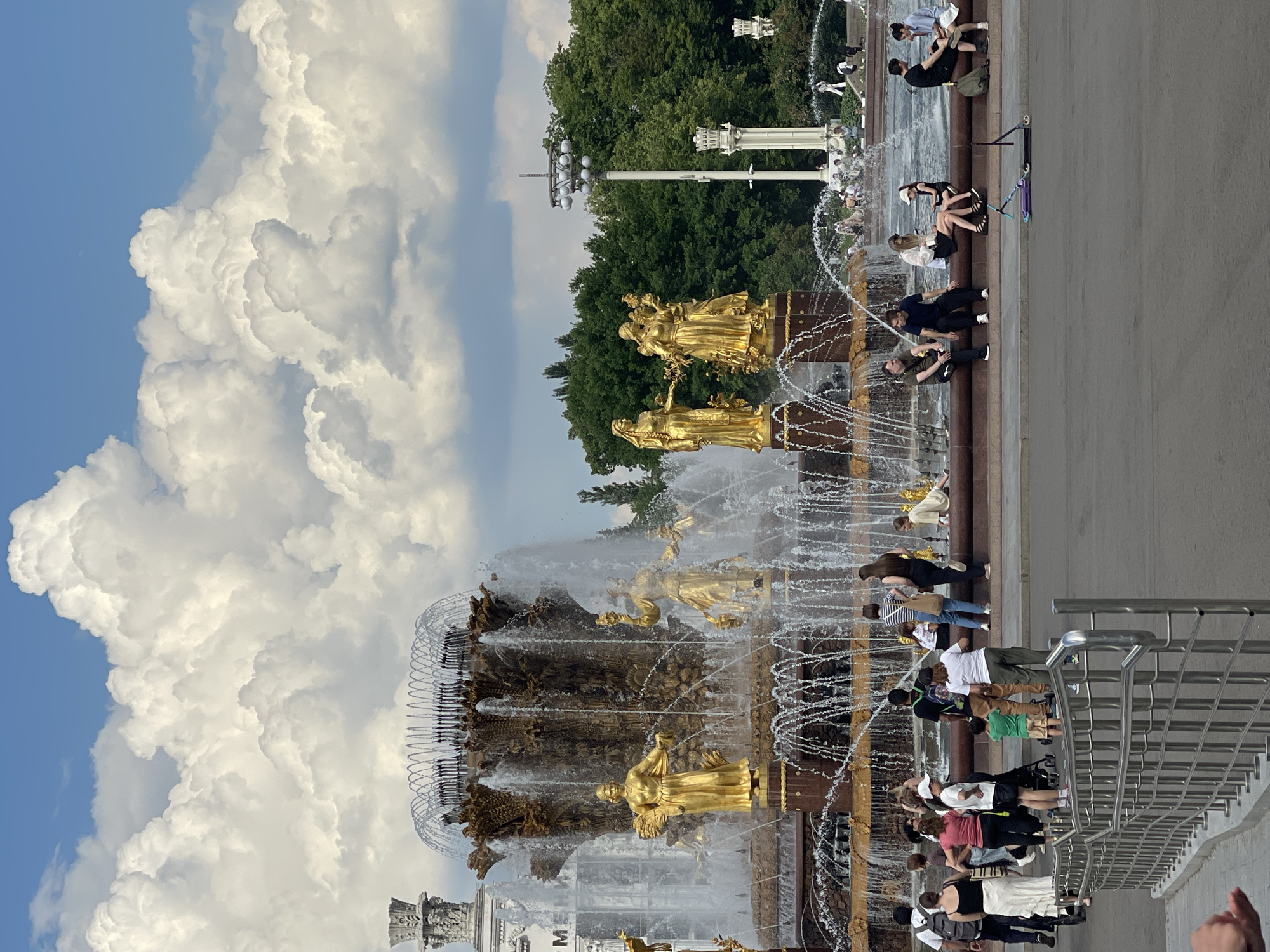 The image depicts a sunny day with large, fluffy white clouds in a bright blue sky. In the foreground, a large, ornate fountain is the central focus. The fountain features multiple tiers with golden statues of women holding various items, surrounded by cascading water. People are visible around the fountain, some standing, some sitting on the edge of the fountain's basin, and others walking. Several individuals are dressed in casual summer attire. To the right of the fountain, there are tall lampposts and lush green trees forming the background. The ground in front of the fountain is a paved area where a few scooters are parked. A metal railing is present in the lower left corner of the image.