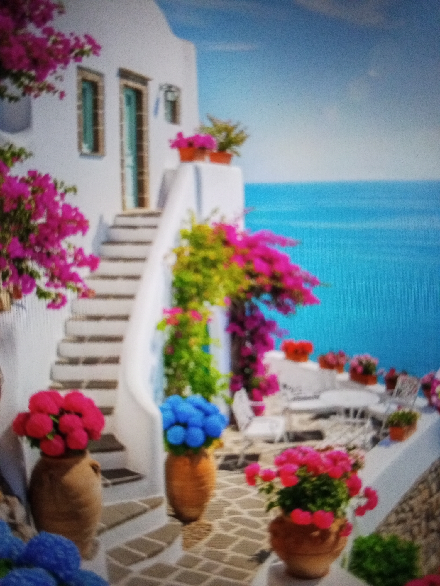 The image depicts a scene reminiscent of the Greek Islands, possibly Santorini, but **not** the city of Tinaquillo, Venezuela. The architecture features white buildings with blue accents. A staircase ascends along the side of one building, adorned with pink bougainvillea. Numerous potted plants with pink and blue flowers are scattered throughout the scene. In the background is a view of the sea. Tables and chairs suggest an outdoor dining or relaxation area. The scene conveys a serene, picturesque atmosphere. There is no evidence of the location being Tinaquillo, Venezuela.