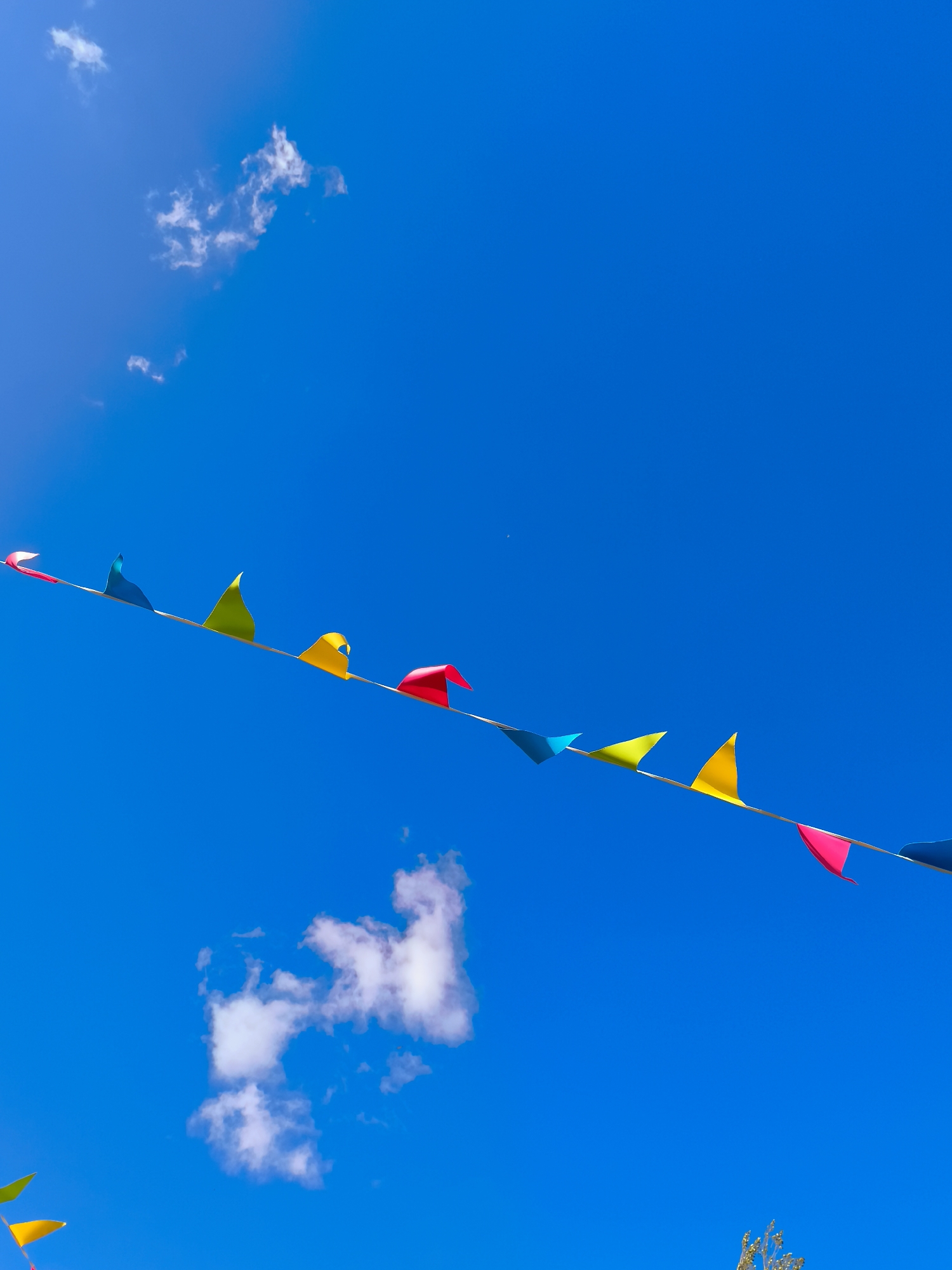 The image shows a string of colorful triangular flags stretched across a bright blue sky. There are a few wispy white clouds scattered in the sky. The flags are predominantly in shades of pink, yellow, blue, and green, and they appear to be made of lightweight material, fluttering in the wind. The string of flags is angled diagonally from the lower left to the upper right of the frame. The scene suggests an outdoor celebration or event taking place on a clear day. This image does not contain any information that would allow for a specific location within Gdańsk, Poland to be identified.