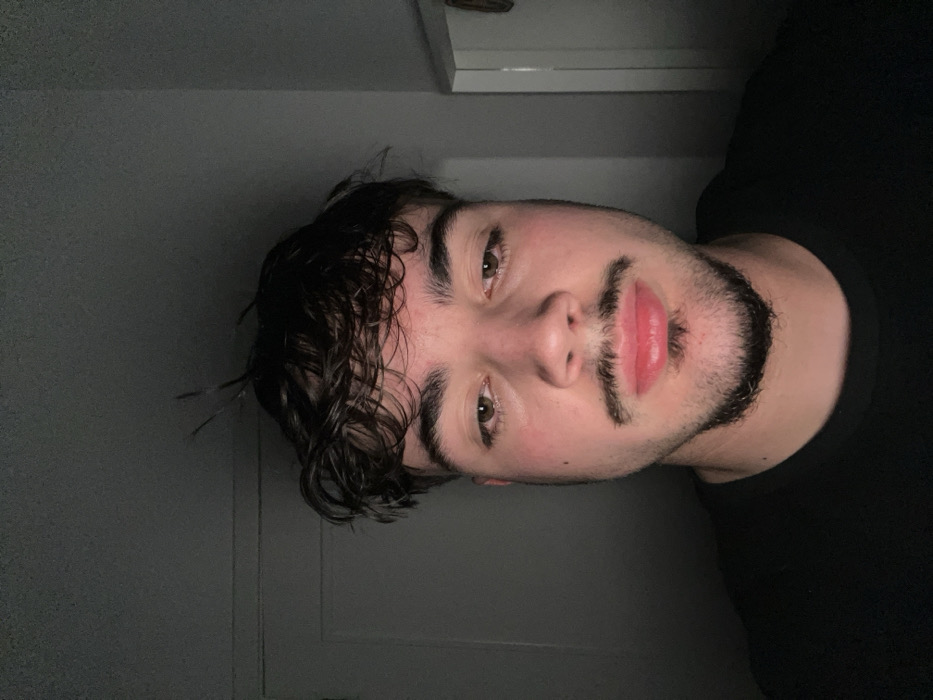 The image presents a close-up, chest-up portrait of a male individual, observed indoors in Vila Real, Portugal. The subject, appearing to be a young adult, directly faces the camera with a neutral expression. He has dark, wavy hair, light brown eyes, a dark mustache, and a trimmed goatee extending to his chin. His lips are full and appear pinkish-red, with the lower lip exhibiting minor swelling or chapping. The subject is wearing a plain, dark-colored crew-neck shirt. Illumination emanates primarily from a frontal-low source, casting distinct upward shadows beneath facial features such as the nose and chin. The background is a dimly lit, indistinct interior, featuring subtle vertical architectural elements consistent with light-colored door frames on the left and right sides of the subject.