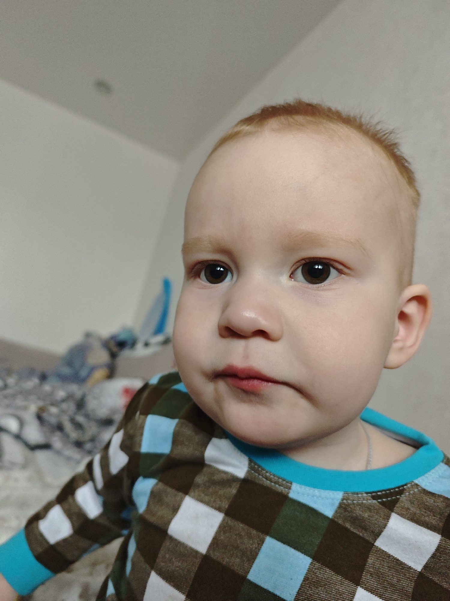 A close-up shot shows a toddler with light skin, red hair, and brown eyes. The child is wearing a long-sleeved shirt with a blue and brown checkered pattern and a blue collar. A silver necklace is visible around the child's neck. The background appears to be an interior space, with a bed and a white wall partially visible. The location is Markovskii, Russia.