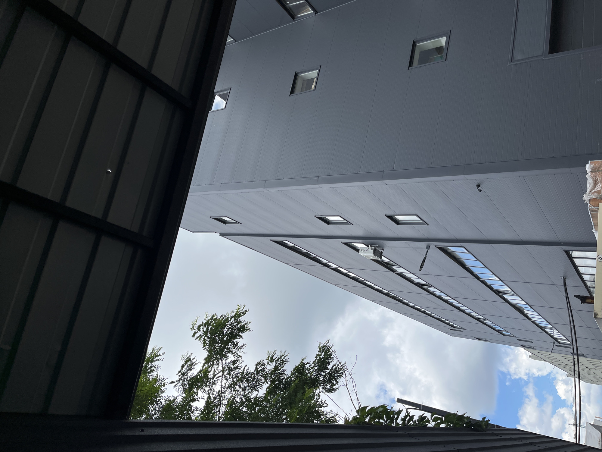 The image shows an upward view of a modern, gray, multi-story building with rows of rectangular windows. The building's exterior appears to be constructed of corrugated metal panels. In the foreground, a dark, corrugated metal overhang or roof is visible, creating a framing effect. Green trees with dense foliage are seen below the building, peeking through the gap between the foreground structure and the building itself. The sky above is cloudy with patches of blue, suggesting daytime. The scene captures an architectural detail with a touch of nature, possibly in an industrial or commercial area. There is no visible text in the image. The perspective is dramatic, emphasizing the scale of the building against the sky and trees.
