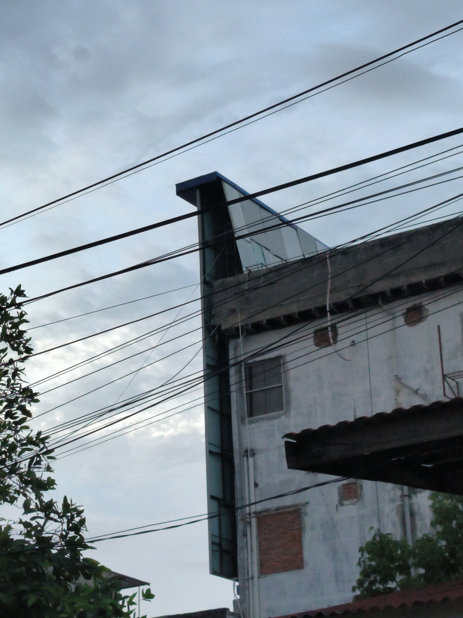 An eye-level, outdoor shot depicts a building, possibly in Lhokseumawe, Indonesia, under a cloudy sky. The building features both old and modern architectural elements: a concrete facade with bricked-over windows sits alongside a newer, glass-encased section that extends upwards. Overhead electrical wires are strung haphazardly across the scene, partially obscuring the view. The bottom of the frame contains foliage from nearby trees and a partial view of a red tile roof.