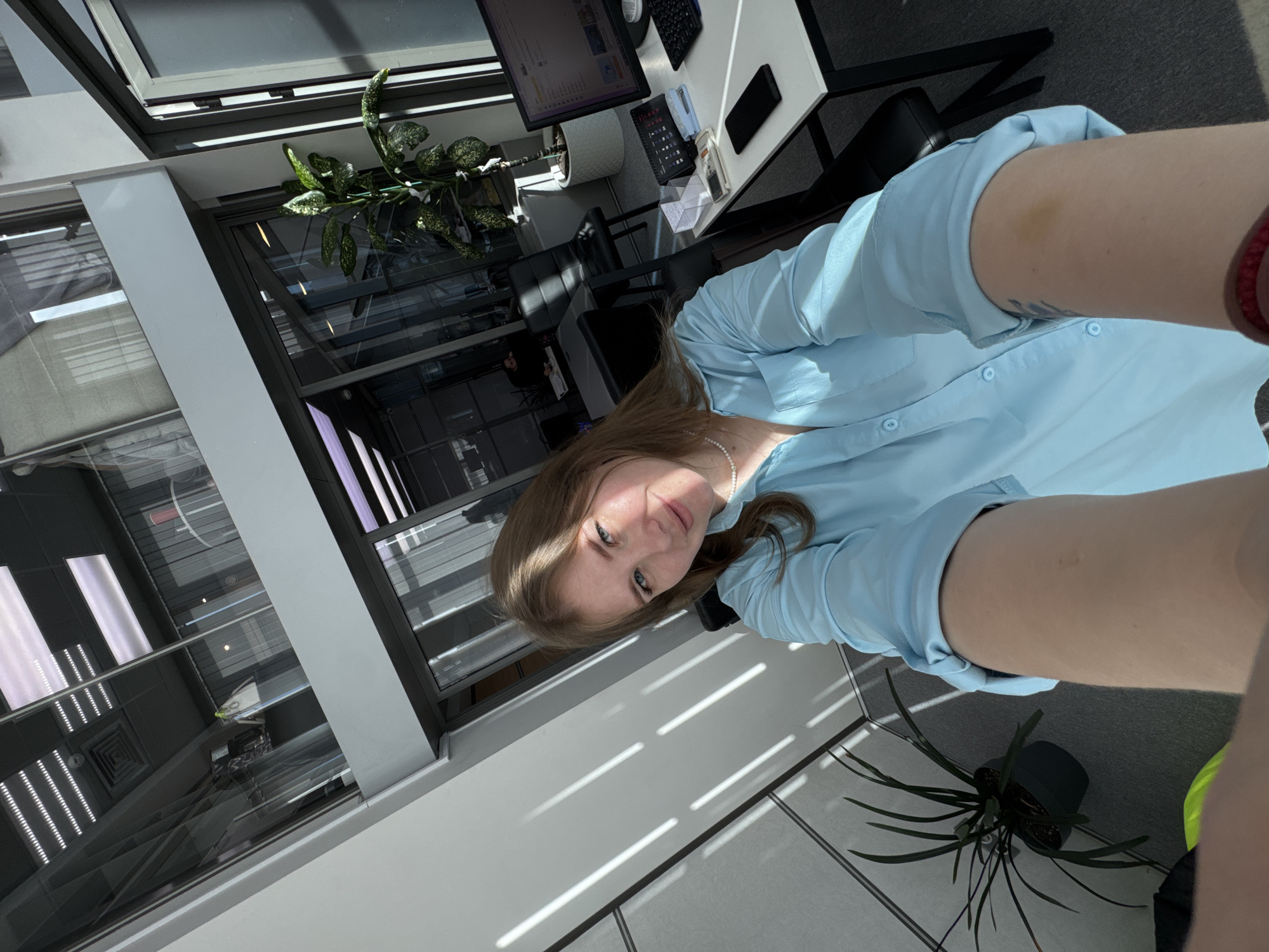 The image shows a woman with fair skin and long brown hair, wearing a light blue button-down shirt and a white pearl necklace. She is indoors, seemingly in an office environment. A desk with a computer, keyboard, and other office supplies is visible in the background. A plant in a white pot sits on the desk. Large windows with a view of the city reflect overhead lights and possibly buildings. Another plant is on the floor next to the windows. The image is angled as if taken in a selfie style.
Based on the visible architecture, the location might be a modern office building in Perm, Russia, given the presence of similar structures in the city.
