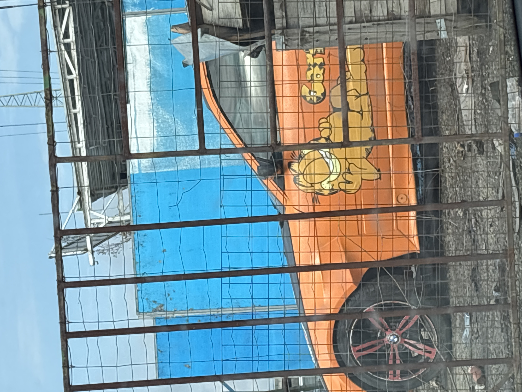 The image shows a fenced-off area in Burgas, Bulgaria, with an orange vehicle visible behind the fence. The vehicle has a cartoon graphic of Garfield on its side. The tire of the car features red accents. Behind the car is a building with blue panels. The fence is made of metal grids and bars. The background is slightly blurry, but appears to include vegetation and other objects.