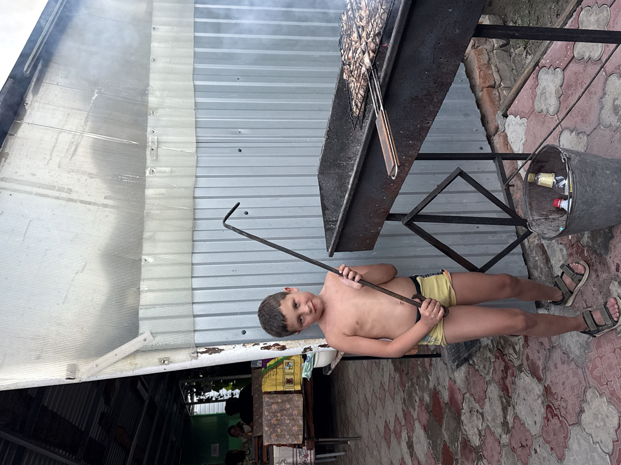 A young boy, shirtless and wearing yellow shorts and sandals, stands outdoors. He is holding a long metal skewer over a grill where food is cooking, producing smoke. The boy is smiling at the camera. Behind him, a corrugated metal structure serves as a wall. The ground is paved with decorative tiles. A metal bucket sits nearby, containing several bottles. The scene appears to be set in a backyard or outdoor cooking area, possibly in Sol-Iletsk, Russia. The lighting suggests it is daytime.