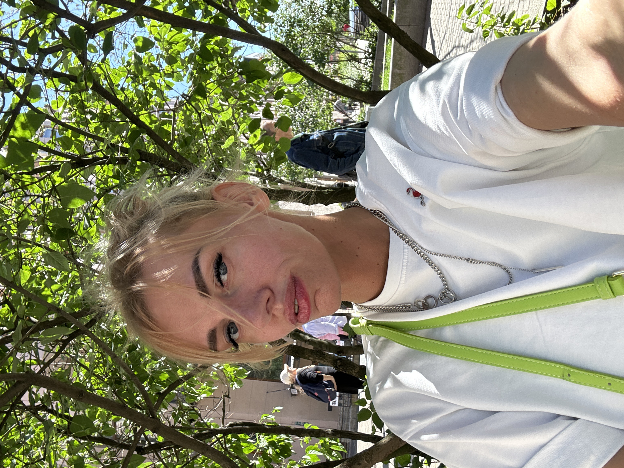 A young woman with blonde hair and blue eyes is taking a selfie outdoors. She is wearing a white t-shirt, a lime green purse strap, and a silver chain necklace. Her hair is styled in a messy bun, and she is looking directly at the camera with her lips pursed. In the background, there are trees with green leaves, a building, and other people walking. The lighting suggests it is daytime and sunny. The presence of Cyrillic text on a sign behind one of the individuals in the background, specifically the word "ФОТО," indicates the location is Saint Petersburg, Russia.