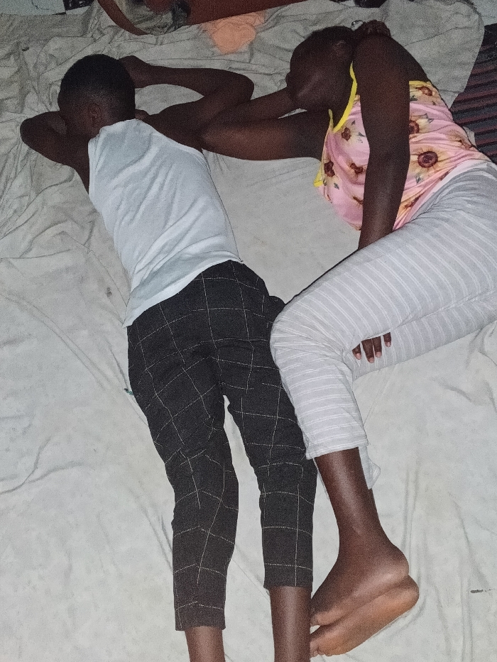 Two individuals, one appearing male and one appearing female, are positioned on a light-colored, wrinkled bed sheet. The male-presenting individual is on the left, lying prone with their head turned away from the camera, wearing a white tank top and dark, grid-patterned trousers. Their arms are bent, with hands near their head. The female-presenting individual is on the right, lying on their left side facing the first individual, wearing a pink and yellow floral sleeveless top and light grey striped leggings. Their bare feet are visible at the bottom right. The female-presenting individual's right arm extends towards the first individual's back. Both individuals appear to be sleeping or resting. The location is an indoor setting in the city of Unknown, Unknown.