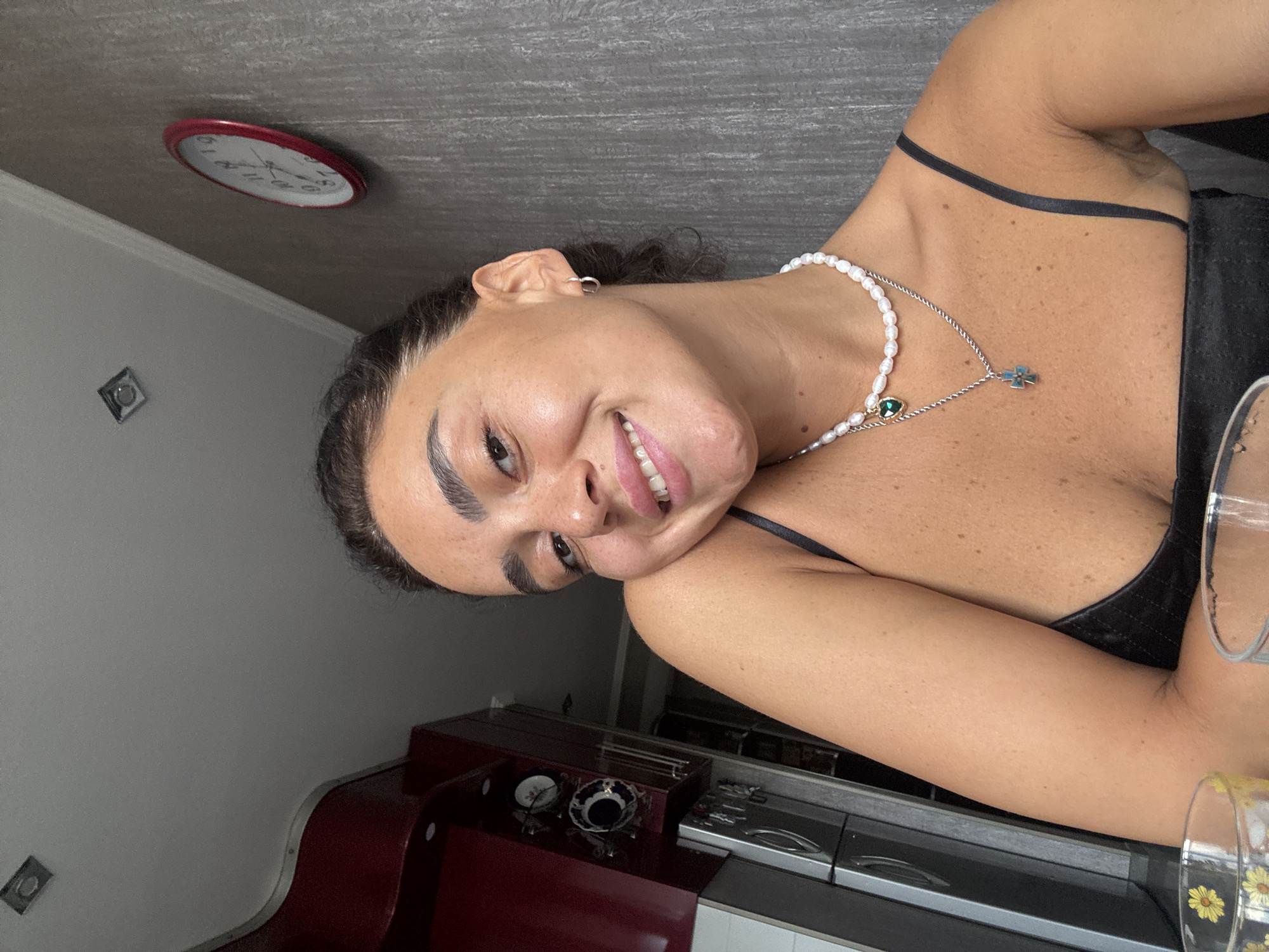 A medium shot captures a woman in her early 20s, with dark hair pulled back, looking directly at the camera with a wide smile. Her skin is lightly tanned and freckled, and she wears a black spaghetti strap top, a pearl necklace, and a silver chain with a cross pendant. She has a small hoop earring in her right ear. In the background, a red-faced clock is mounted on a textured gray wall, and a dark red cabinet with a mirrored surface is visible to the left. The scene appears to be set indoors, possibly in a kitchen or dining area.