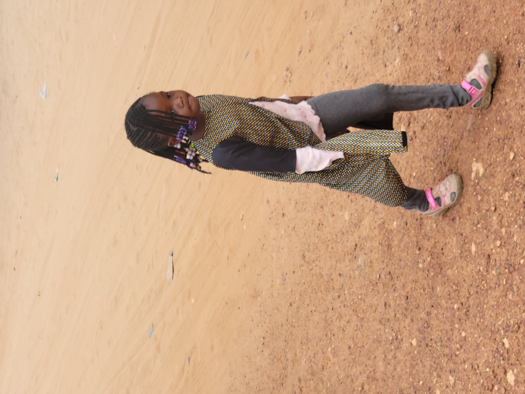 An African girl is seen on the ground in Gura Top, Nigeria. She is wearing gray leggings, a black and yellow patterned long-sleeved jacket, and pink sandals. Her hair is braided with colorful beads. The ground appears to be dirt and sand.