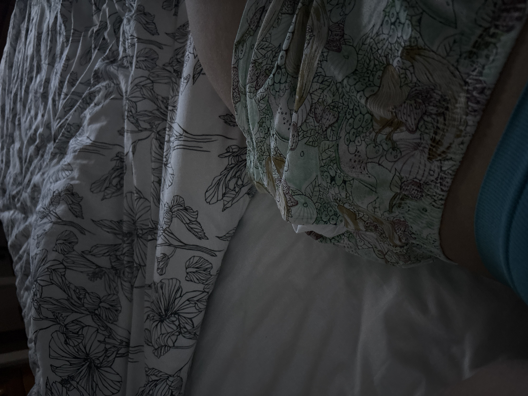 The image displays an indoor scene in Kyustendil, Bulgaria, featuring a bed viewed from an overhead perspective. The bed is covered by a duvet with a white background and a black-and-white line art floral pattern depicting large blossoms and leaves. Beneath the duvet, a plain, light-colored sheet is partially visible. A portion of a person's upper thigh and lower torso is discernible in the lower right area of the frame. The individual is wearing sleepwear, consisting of patterned shorts or pajama bottoms. This garment is light mint green with an intricate motif of shells, coral, and abstract marine life rendered in muted brown, purple, and white outlines, displaying a ruffled or elasticated waistband. Below this patterned garment, a solid teal-colored fabric is visible. The scene is dimly lit.