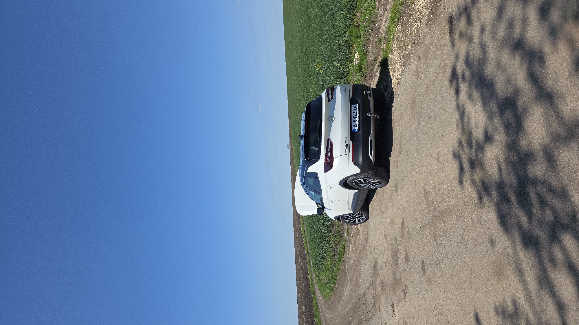 A white Citroen C4 Cactus car, license plate B 8922 XK, is parked on a rural dirt road next to a green field. The location is likely near Dobrogled, Bulgaria. The sky is blue and clear. Tree shadows are visible on the road.