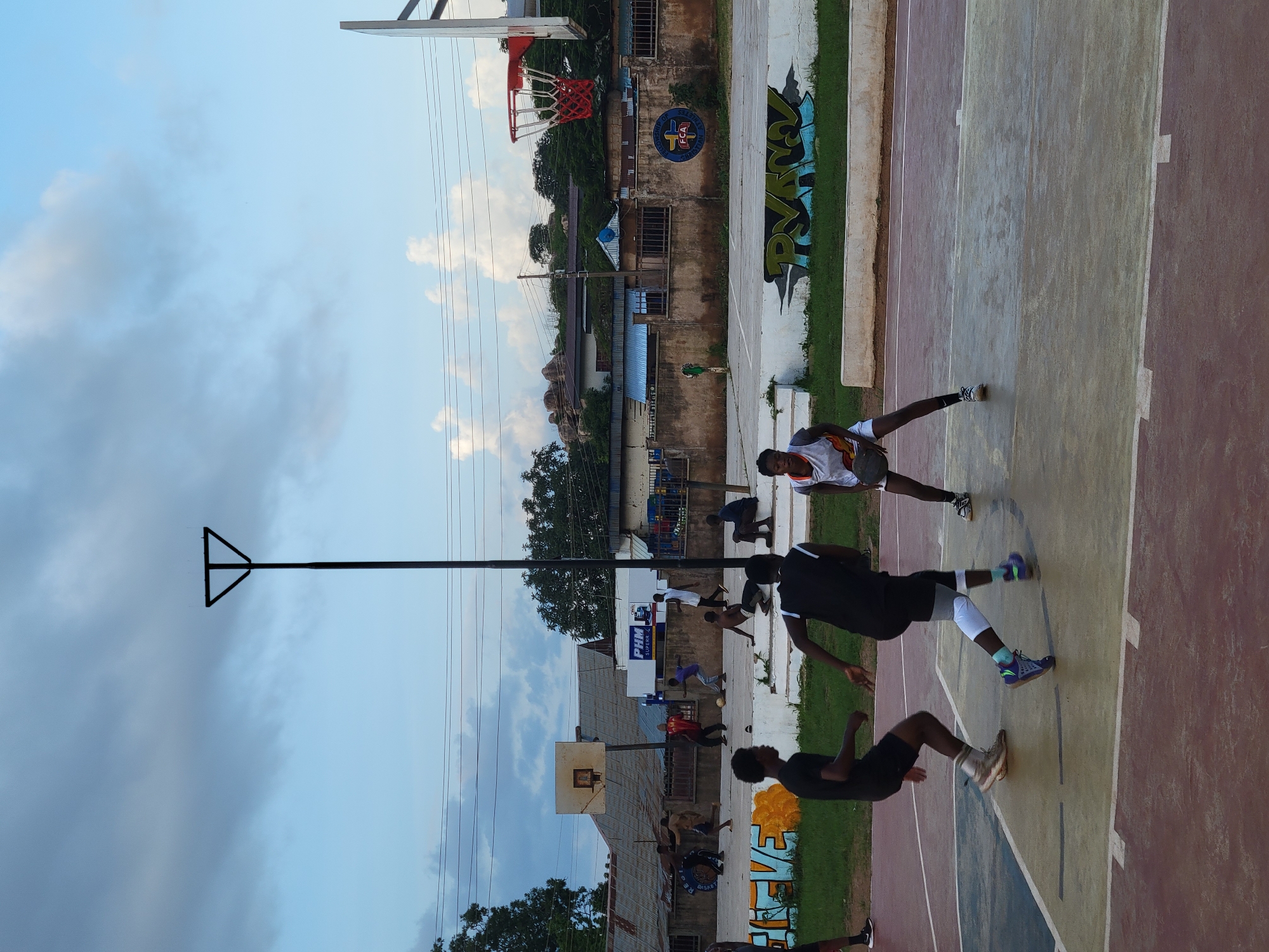 The image depicts an outdoor basketball court in Jos, Nigeria. Several individuals, primarily young men and boys, are engaged in a basketball game. One person in the foreground, wearing a white and orange basketball jersey and shorts, is holding a basketball. Other players are in various stages of movement, dribbling, defending, or preparing to shoot. The court is made of colored asphalt with white boundary lines. In the background, there are buildings with some graffiti on the walls, a basketball hoop with a red net, and a basketball pole. The sky is overcast with soft clouds, suggesting it might be late afternoon or early evening. The overall atmosphere appears energetic and focused on the game. There is some graffiti on the wall, and a sign with "PHM SUPER C" is visible.