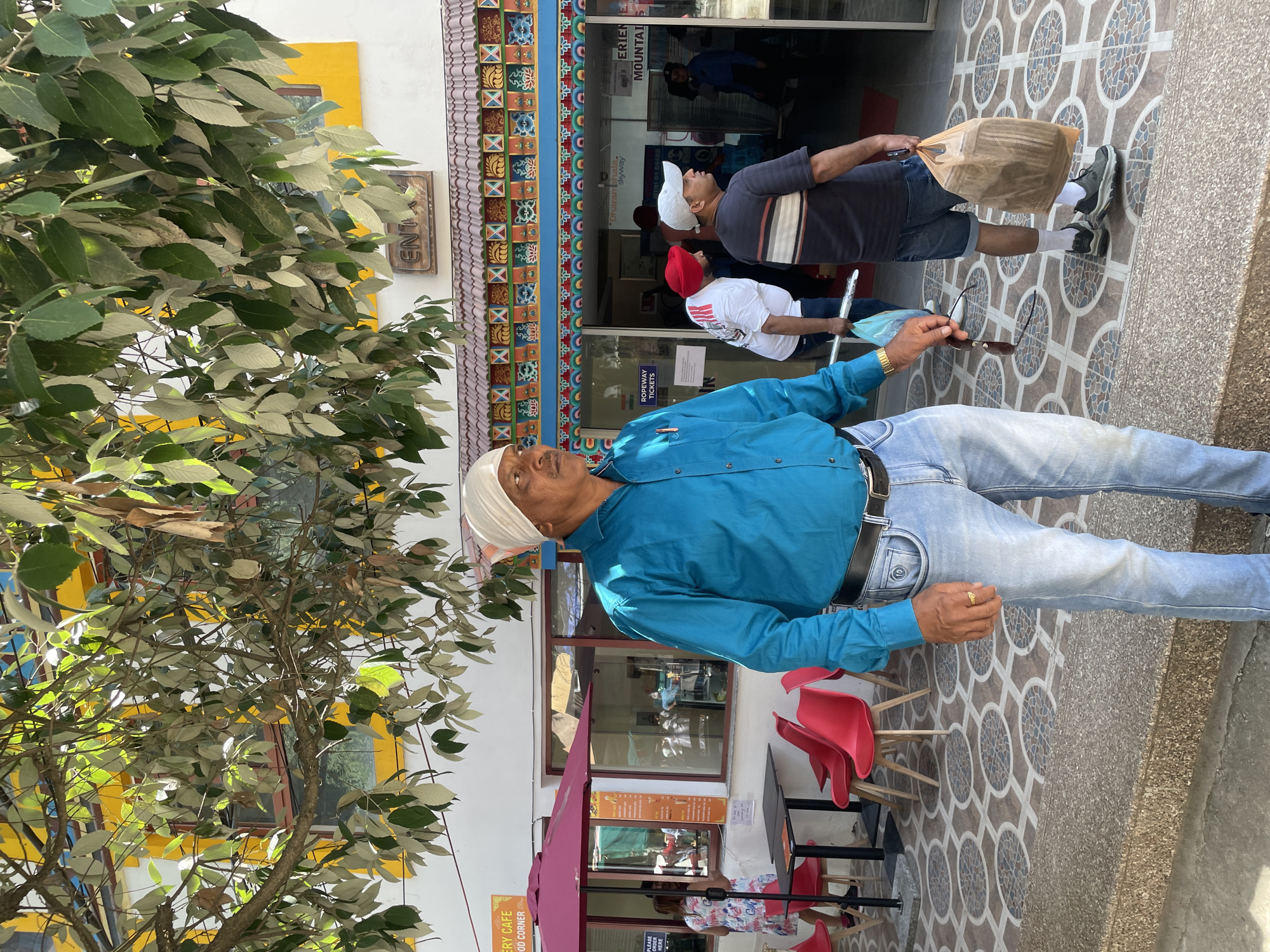 The image shows a street scene in Dharamshala, India. An older man wearing a white head covering, a blue shirt, and light-wash jeans stands in the foreground. He is looking up and holding sunglasses. Behind him, two men walk toward the entrance of "Eriene Mountain Skyway," where signs mention "Ropeway Tickets" and "Dharamshala Skyway." One man wears a white cap and a dark striped shirt, carrying a brown bag. The other man has a red cap and a white shirt.
A cafe is located to the left, with red chairs and a pink umbrella. A tree is visible on the left side of the image. The building has a colorful border design above the entrance and a yellow wall section. The sidewalk has a patterned tile design.