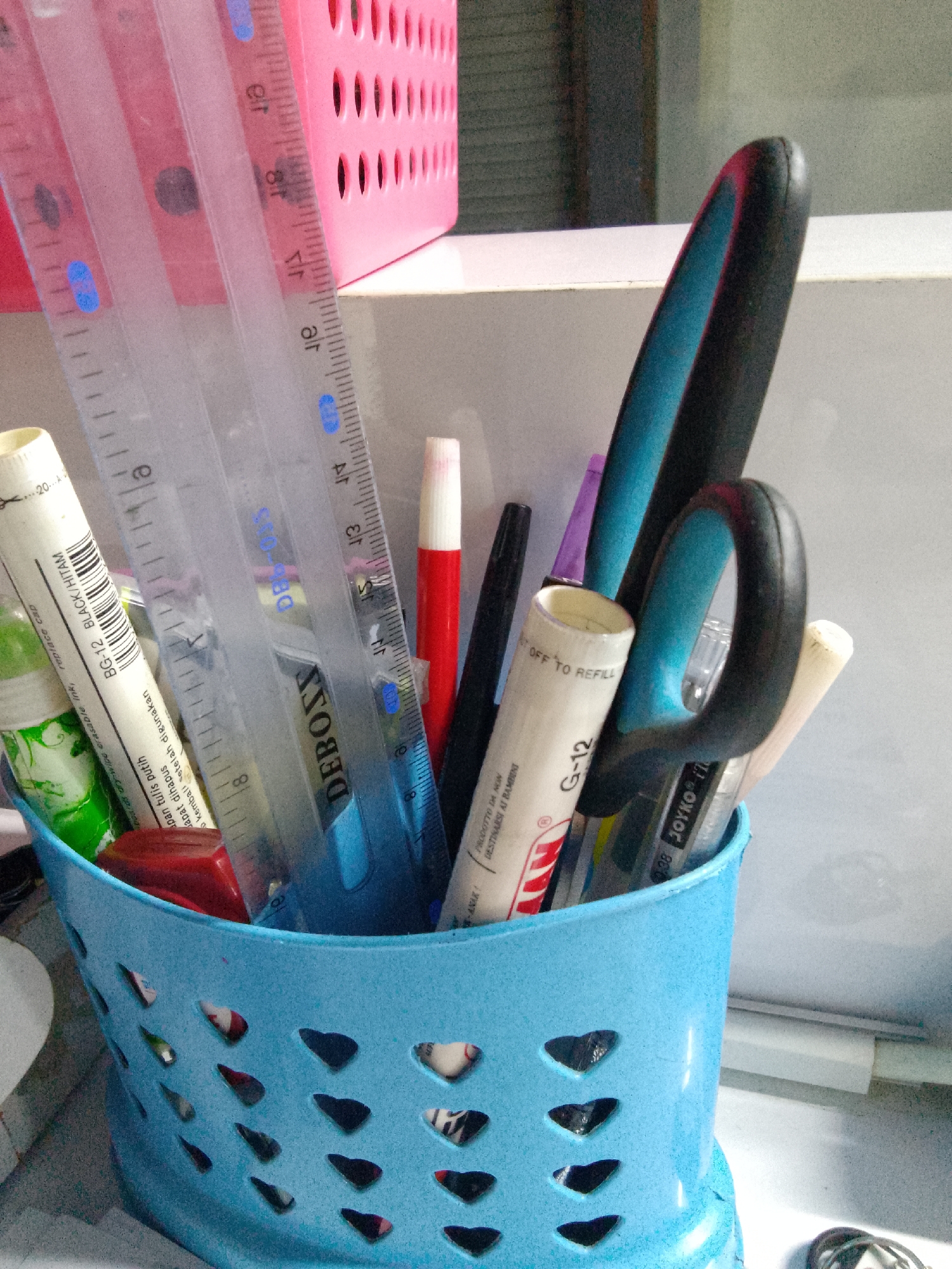 The image depicts a close-up of a blue plastic basket overflowing with various stationery items. The basket is filled with pens, markers, scissors, and rulers, all arranged in a haphazard manner. In the background, a pink plastic container with a perforated design is visible, along with a white surface and a blurred background.
The scene is set in a room, likely a home or office, with a focus on the desk or tabletop where the stationery items are kept. The lighting is natural, coming from a window or overhead source, casting shadows on the objects and highlighting their textures.
No individuals are present in the image, and no actions are being performed. The image is a static representation of the organized chaos of a collection of writing and art supplies.
The location within the city of Trenggalek, Indonesia, is not discernible from the image itself. The image provides no contextual clues related to a specific geographical location within Trenggalek or any other city.