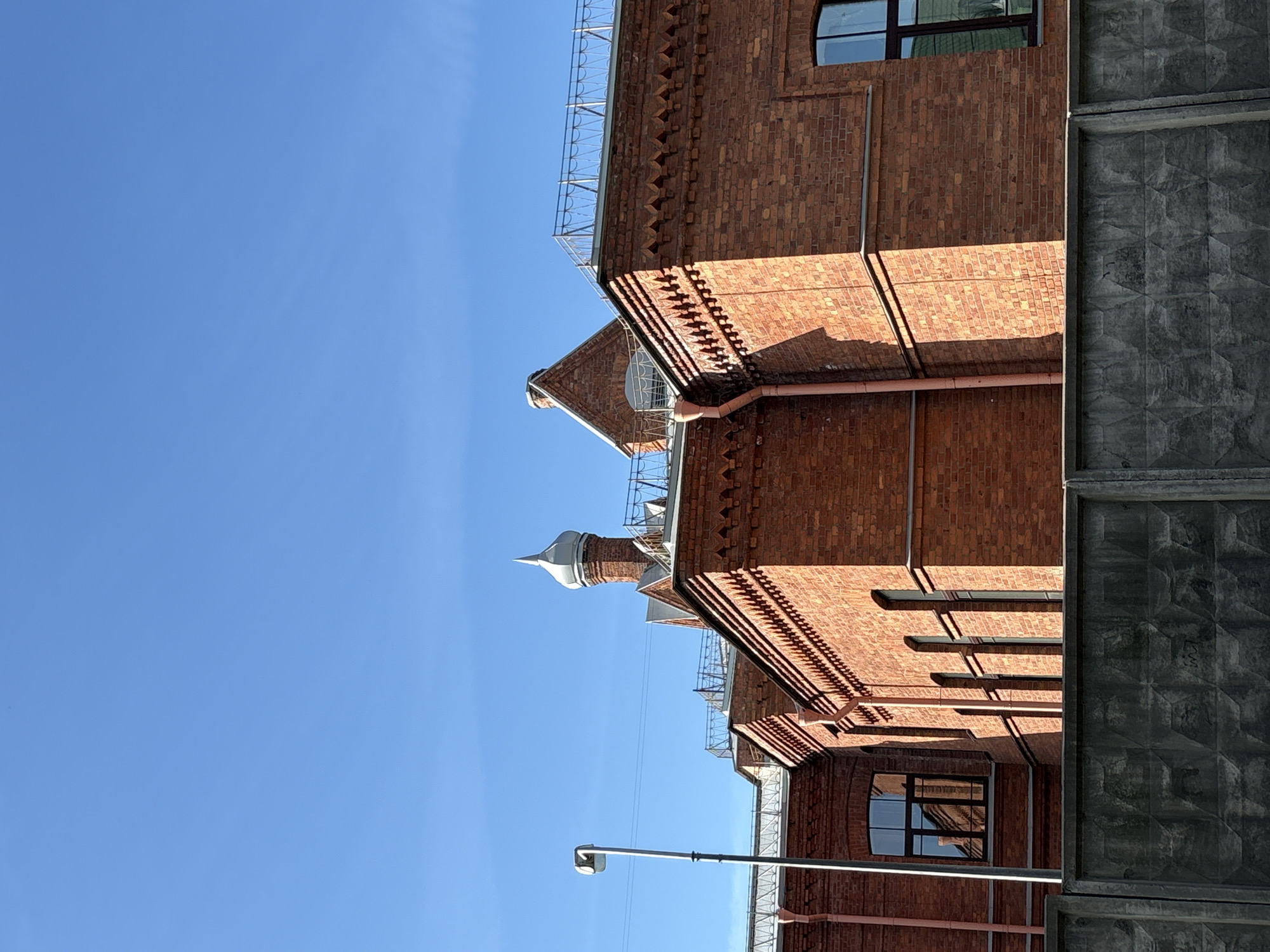 This image captures a vibrant architectural scene, primarily featuring brick buildings against a clear blue sky, under bright sunlight. The perspective is tilted upwards, showcasing the upper sections of the structures.
In the upper portion of the frame, a wide expanse of cloudless, cerulean blue sky dominates, indicating a clear, sunny day. Directly below, two main brick buildings are visible. The building in the foreground and to the right is constructed of reddish-brown brick, exhibiting intricate architectural details. These include decorative friezes with saw-tooth or zigzag patterns running along the eaves and cornices, and multiple dark-framed windows, some rectangular, others semicircular. Metal railings are visible along the rooflines. A light-colored downspout or drainage pipe runs vertically down one section of the wall.
Behind this main building, a smaller, similarly constructed brick structure rises, topped with a distinctive, pointed spire or finial, silvery in color.
To the far right, a tall, grey concrete wall or fence stands vertically, contrasting sharply with the brickwork. This wall features a striking, embossed geometric pattern resembling interlocking cubes or diamonds, adding a textured element to the composition.
In the lower-left section of the image, a grey lamp post with a modern, angular light fixture extends upwards, silhouetted against the sky and part of the brick facade. Strong, crisp shadows are cast across the brick walls and architectural embellishments, highlighting the bright intensity of the sunlight, suggesting it is either morning or afternoon. No people or activities are depicted; the scene is static and focused purely on the urban landscape. No visible text is present.