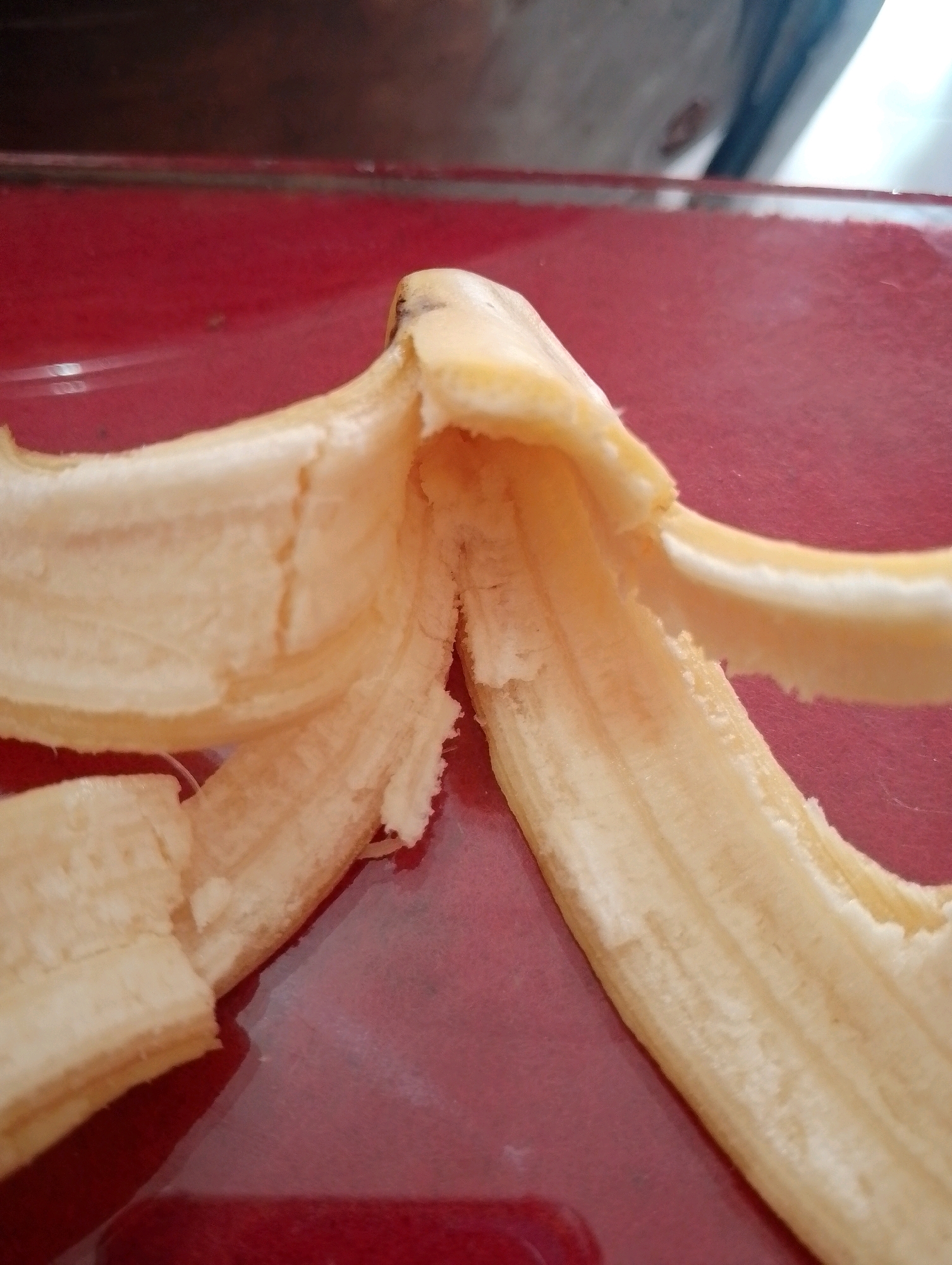 The image shows the inside of a peeled banana, the fruit itself is mostly gone, leaving behind the fibrous peel. The banana peel is light yellow and appears to be slightly moist. It is positioned on a shiny red surface, likely a table or plate, with a dark blurry background. The lighting is bright and comes from above and slightly to the right, creating reflections on the red surface. There are no people or other objects visible in the frame. This image was taken in Papungan, Indonesia.