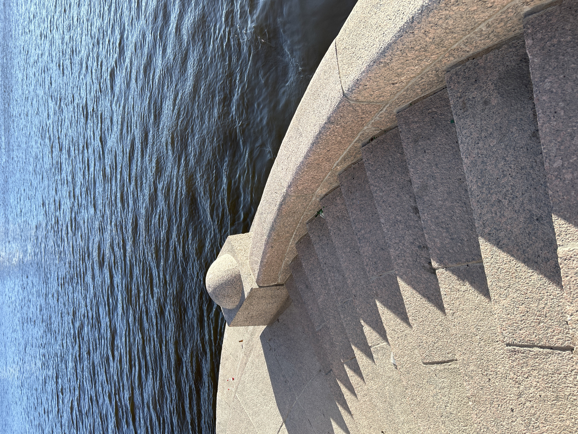 The image presents an elevated, diagonal view of a granite embankment and staircase descending towards a body of water, characteristic of the canals and rivers in Saint Petersburg, Russia. The right side of the frame is dominated by the stone structure, while the left features the water.
The staircase is constructed from speckled grey and pinkish granite, with broad steps casting sharp, triangular shadows onto the flat landing areas. These distinct shadow patterns indicate bright sunlight, likely during the morning or late afternoon. A substantial, curved granite balustrade or railing runs alongside the steps, terminating in a rounded, decorative finial-like element at its base, closer to the water.
The water, which appears dark, displays numerous small ripples and undulations across its surface, reflecting light and suggesting gentle currents or a light breeze. No people are visible, and no specific activity is taking place. The scene conveys a sense of architectural solidity and permanence under clear, sunny weather conditions. There is no visible text.